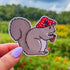 Die cut sticker of a squirrel wearing a red mushroom cap and holding an acorn