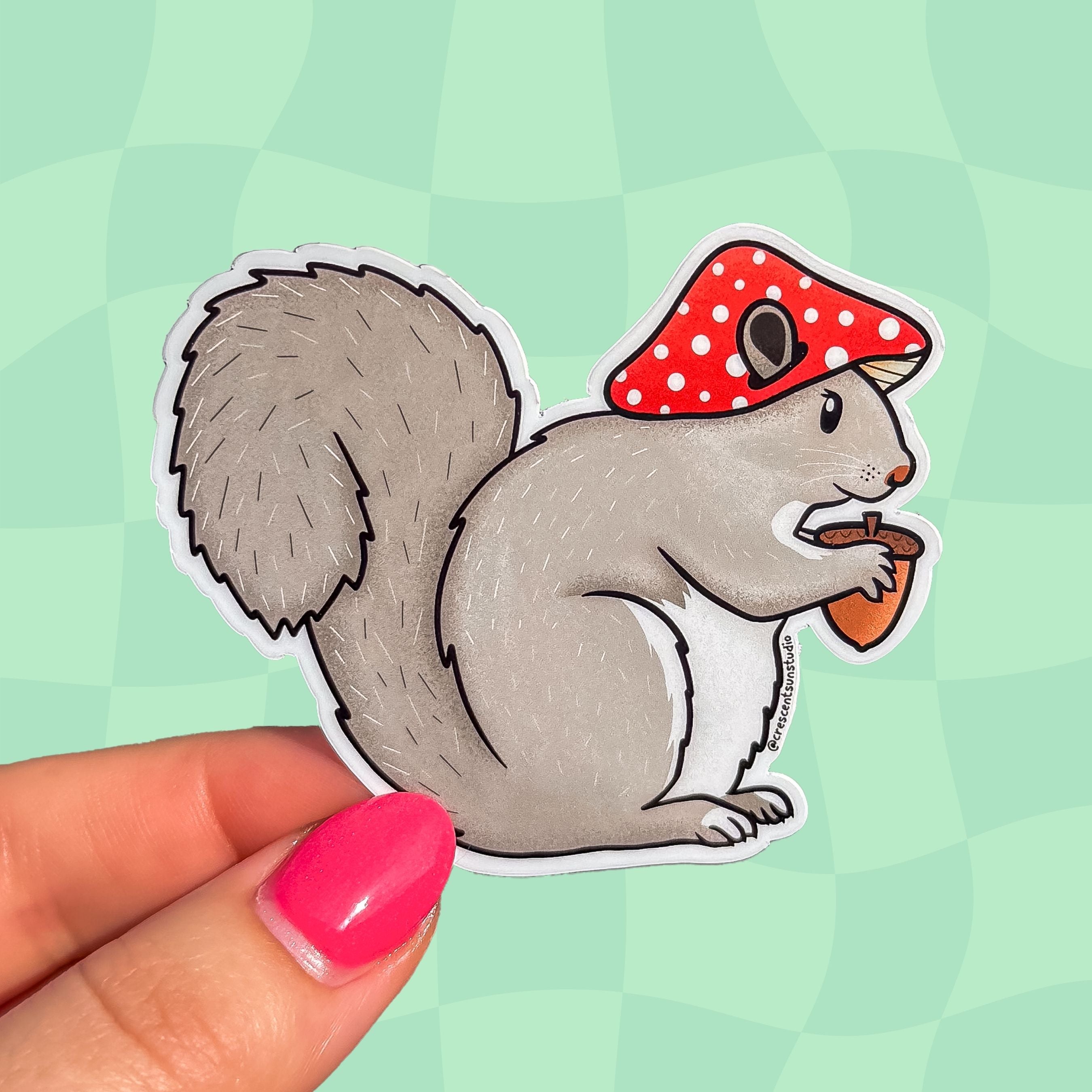 Die cut sticker of a gray squirrel wearing a red mushroom cap and holding an acorn