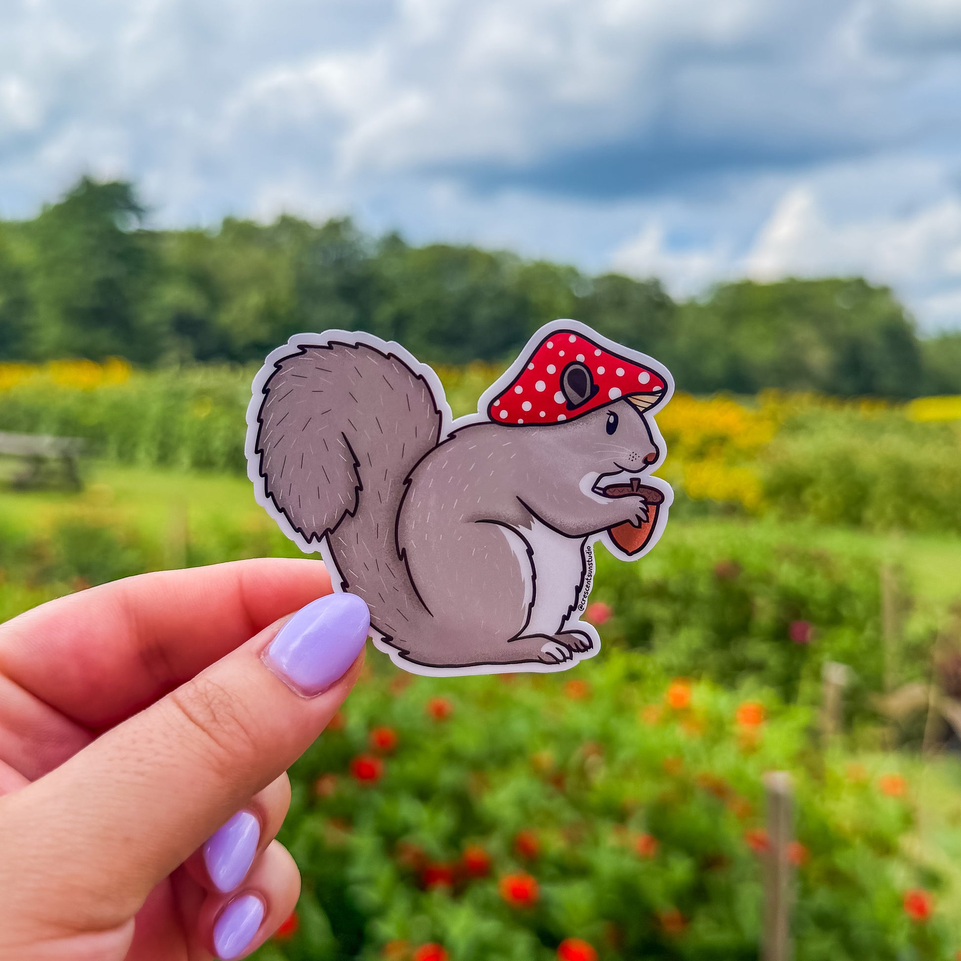 A zoomed out image of a die cut sticker of a squirrel wearing a red mushroom cap and holding an acorn.