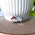 A small enamel pin in the shape of an opossum wearing a red and white mushroom hat. The pin is displayed propped against a large white pot.