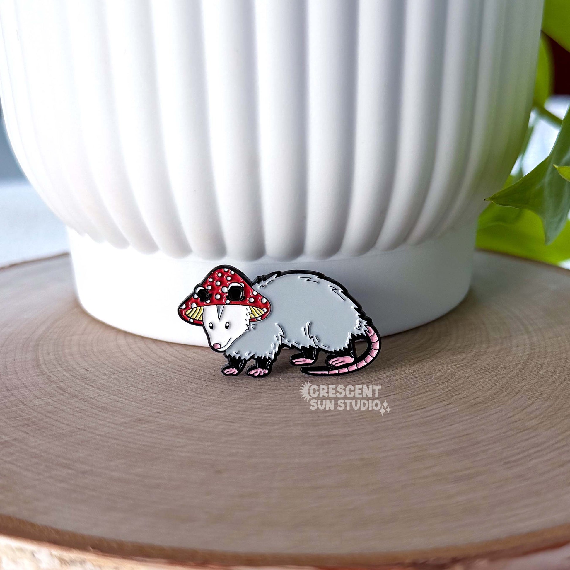 A small enamel pin in the shape of an opossum wearing a red and white mushroom hat. The pin is displayed propped against a large white pot.