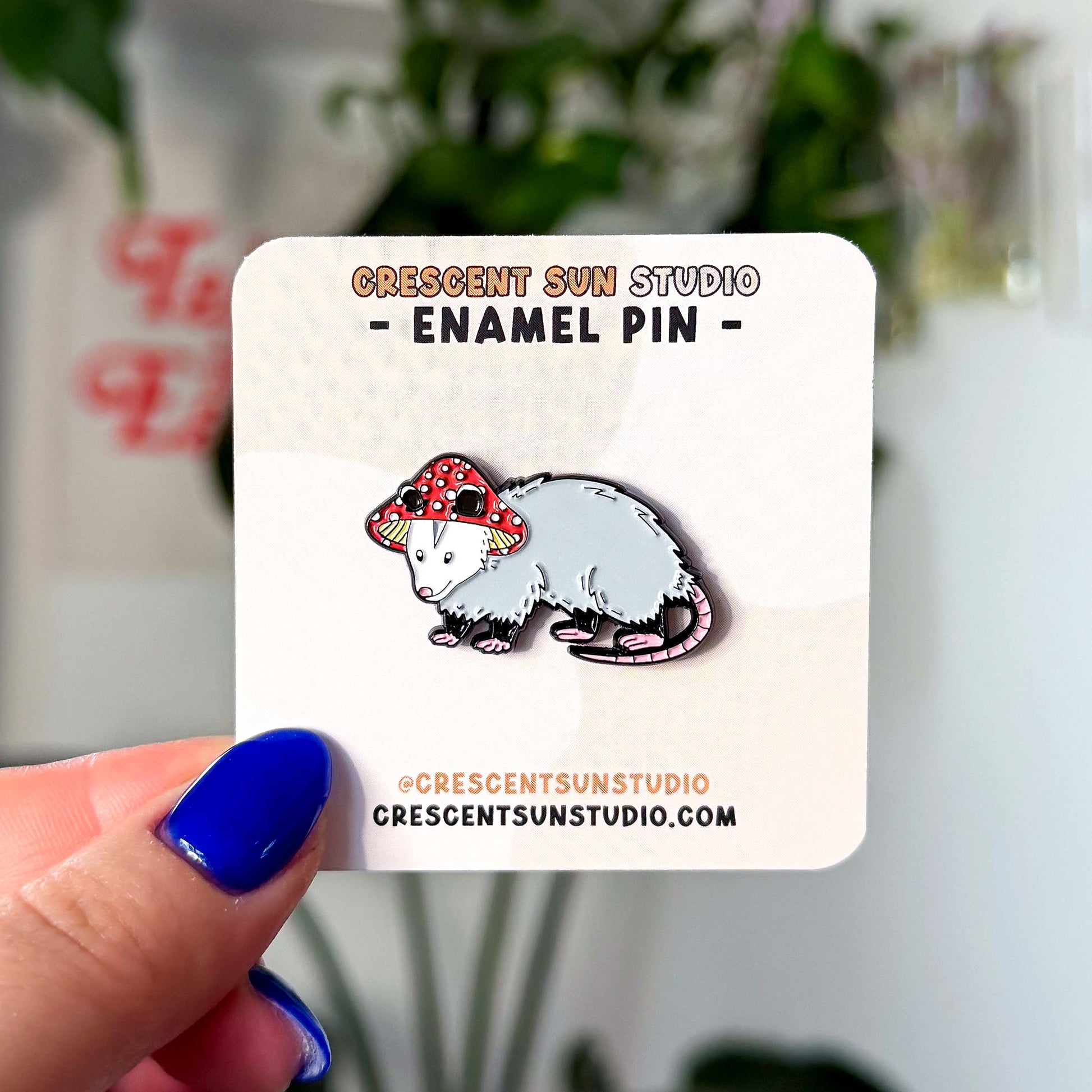 A small enamel pin in the shape of an opossum wearing a red and white mushroom hat. The pin is displayed on a light colored card.