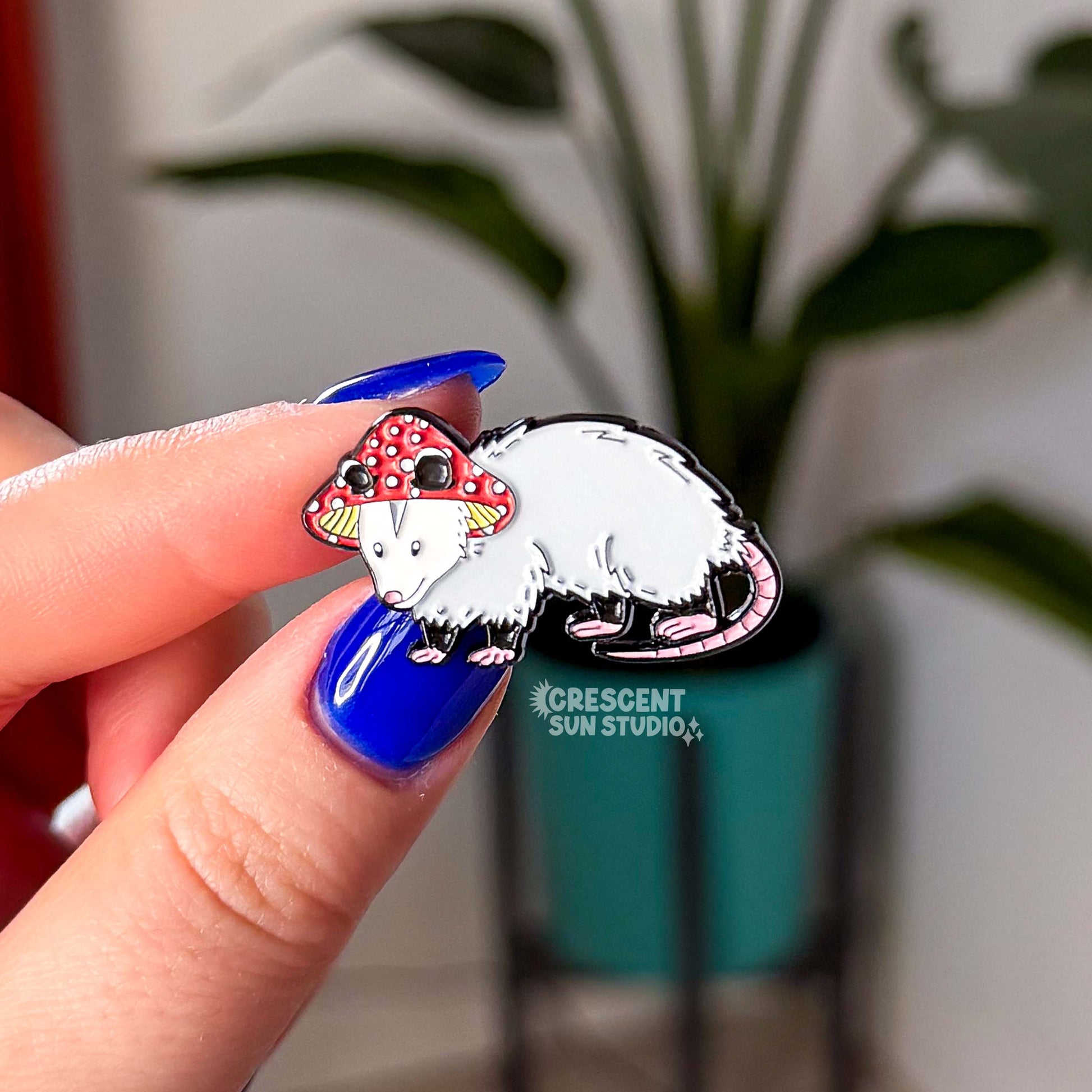 A small enamel pin in the shape of an opossum wearing a red and white mushroom hat. Two fingers with bright blue nail polish hold the pin to the camera.