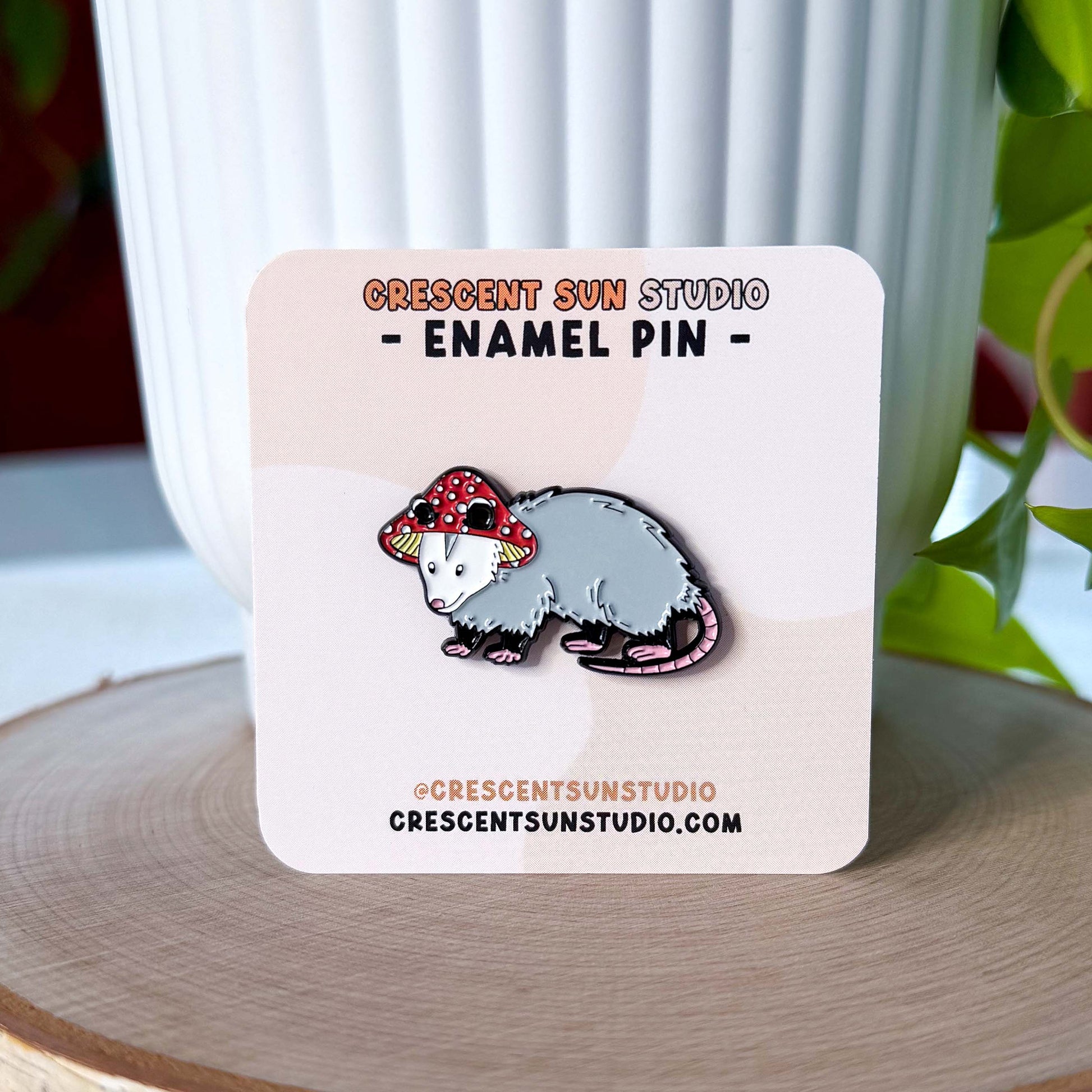 A small enamel pin in the shape of an opossum wearing a red and white mushroom hat. The pin is displayed on a light colored card.