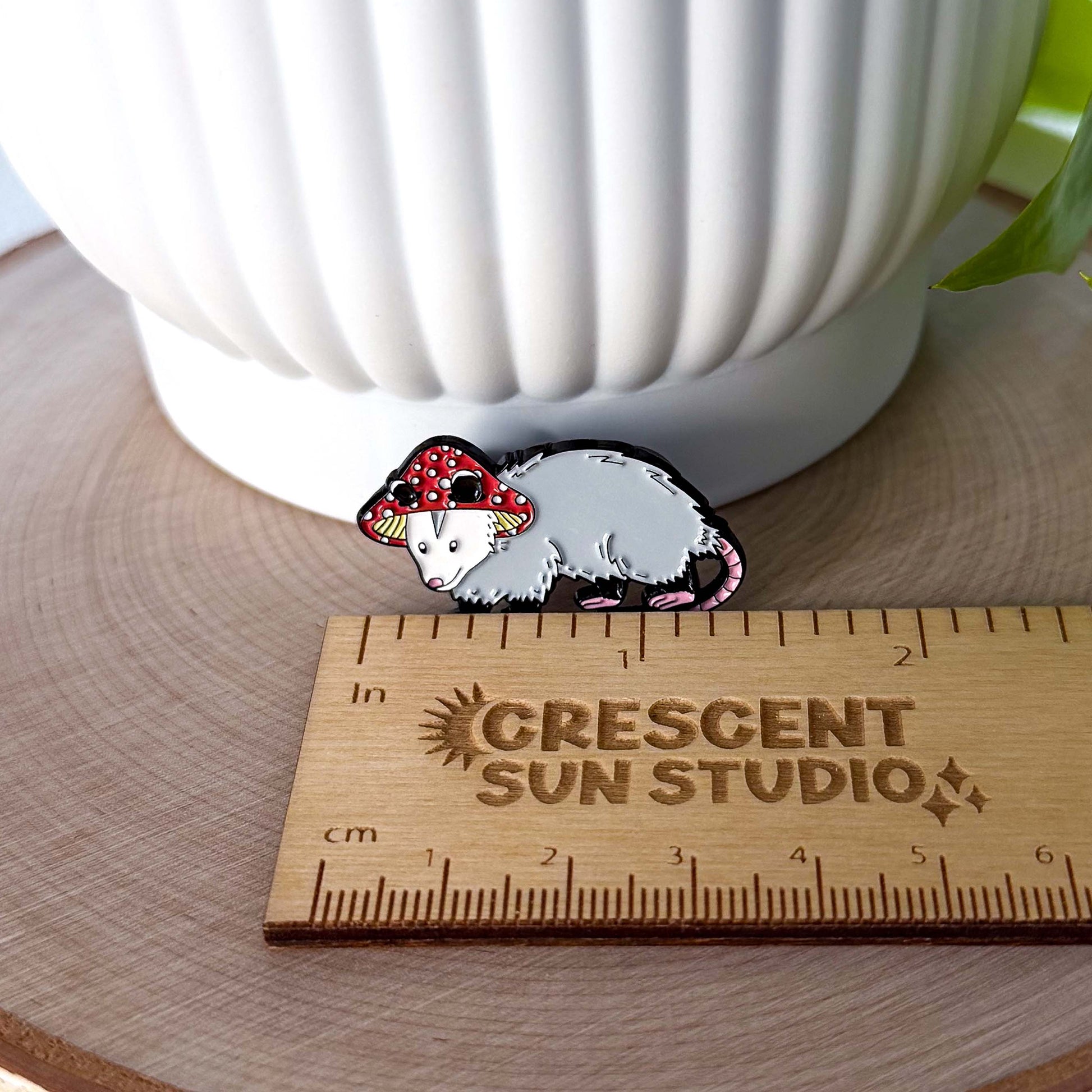 A small enamel pin in the shape of an opossum wearing a red and white mushroom hat. In this image, the pin is next to a ruler showing its width of 1.35 inches.