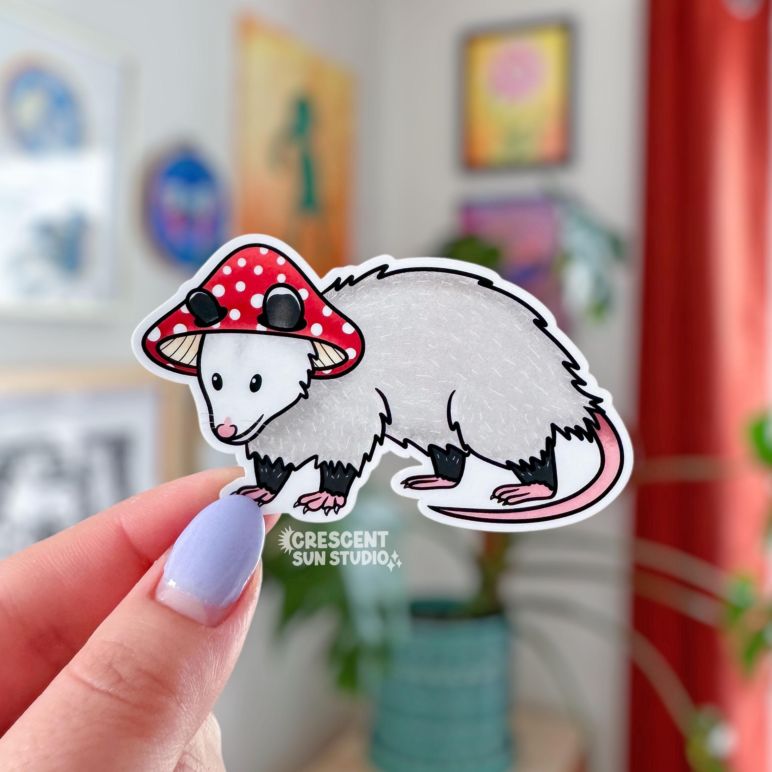 A die cut sticker of an opossum wearing a red and white mushroom cap for a hat.