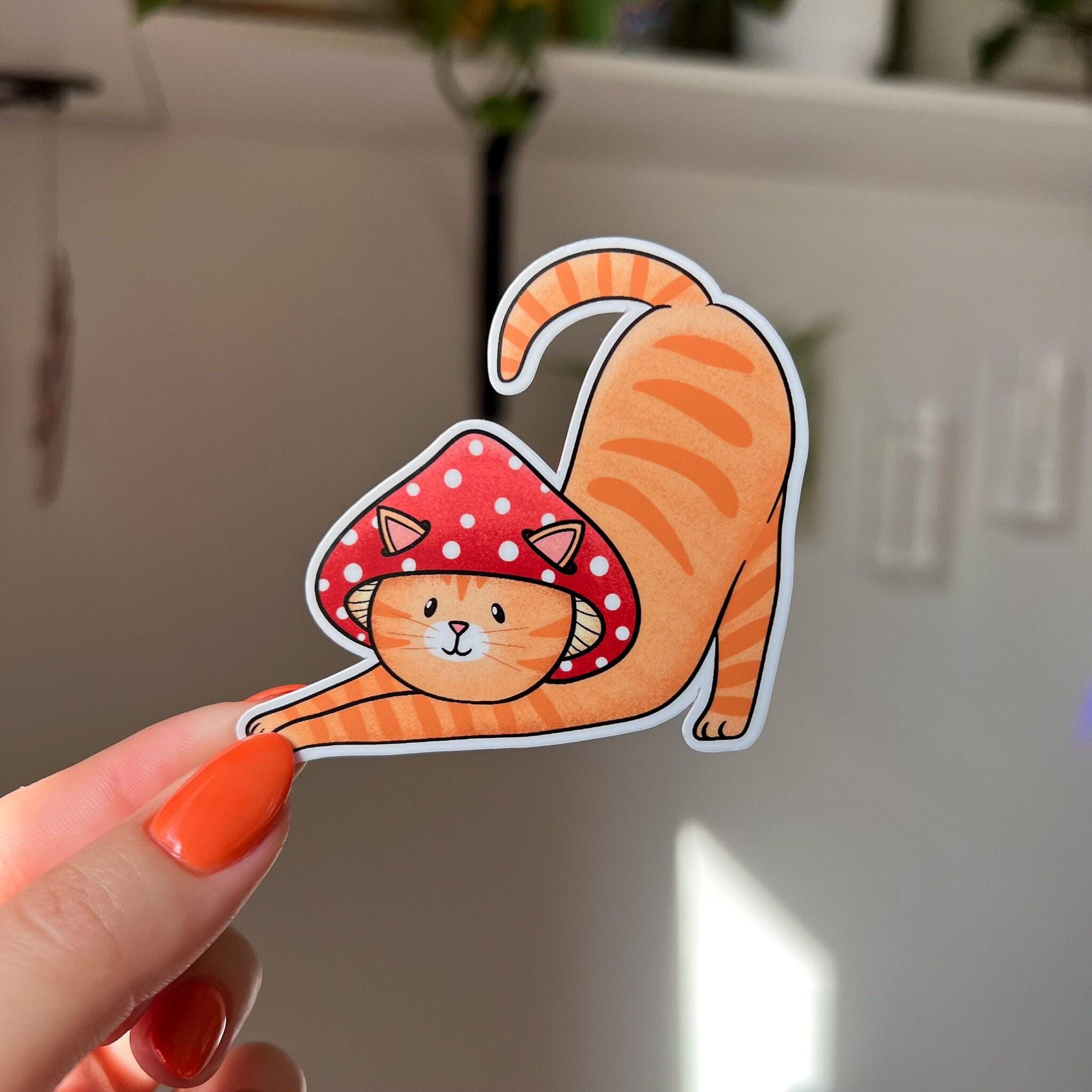 The cat is a tabby (orange with darker orange stripes) and has a sweet expression on his face. His red mushroom hat with white spots has spots for his ears to poke through. The cat is in a stretching pose with his tail curling up behind him.

In this image, the cat-shaped sticker is held close to the camera.