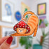 A sticker in the shape of a cat wearing a mushroom hat is held close to the camera. The cat is a tabby (orange with darker orange stripes) and has a sweet expression on his face. His red mushroom hat with white spots has spots for his ears to poke through. The cat is in a stretching pose with his tail curling up behind him.