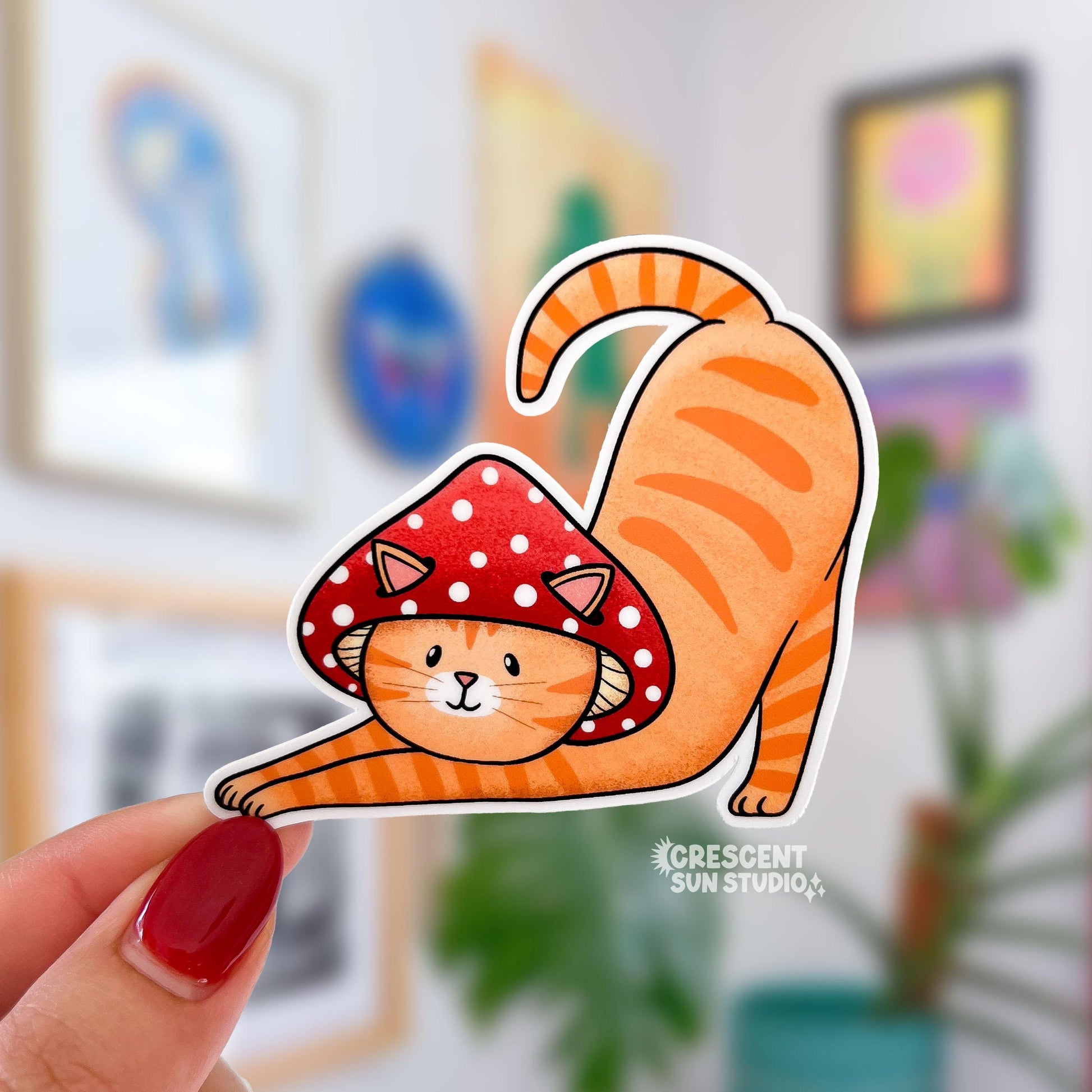 A sticker in the shape of a cat wearing a mushroom hat is held close to the camera. The cat is a tabby (orange with darker orange stripes) and has a sweet expression on his face. His red mushroom hat with white spots has spots for his ears to poke through. The cat is in a stretching pose with his tail curling up behind him.