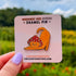 An enamel pin in the shape of a cat wearing a mushroom hat is held close to the camera. The cat is a tabby (orange with darker orange stripes) and has a sweet expression on his face. His red mushroom hat with white spots has spots for his ears to poke through. The cat is in a stretching pose with his tail curling up behind him.