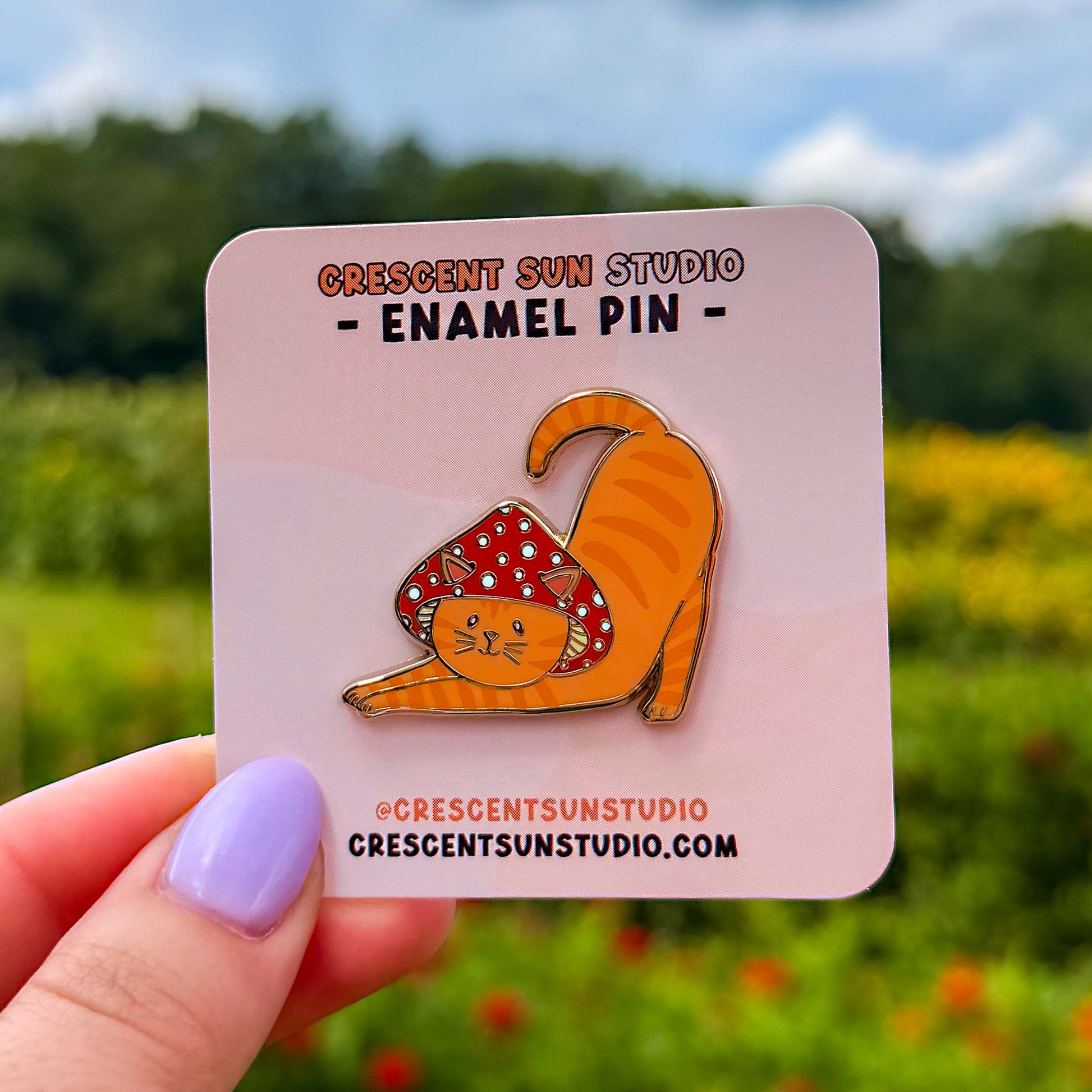 An enamel pin in the shape of a cat wearing a mushroom hat is held close to the camera. The cat is a tabby (orange with darker orange stripes) and has a sweet expression on his face. His red mushroom hat with white spots has spots for his ears to poke through. The cat is in a stretching pose with his tail curling up behind him.