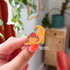 A hand holds an enamel pin in the shape of a cat wearing a mushroom hat is held close to the camera. The cat is a tabby (orange with darker orange stripes) and has a sweet expression on his face. His red mushroom hat with white spots has spots for his ears to poke through. The cat is in a stretching pose with his tail curling up behind him.