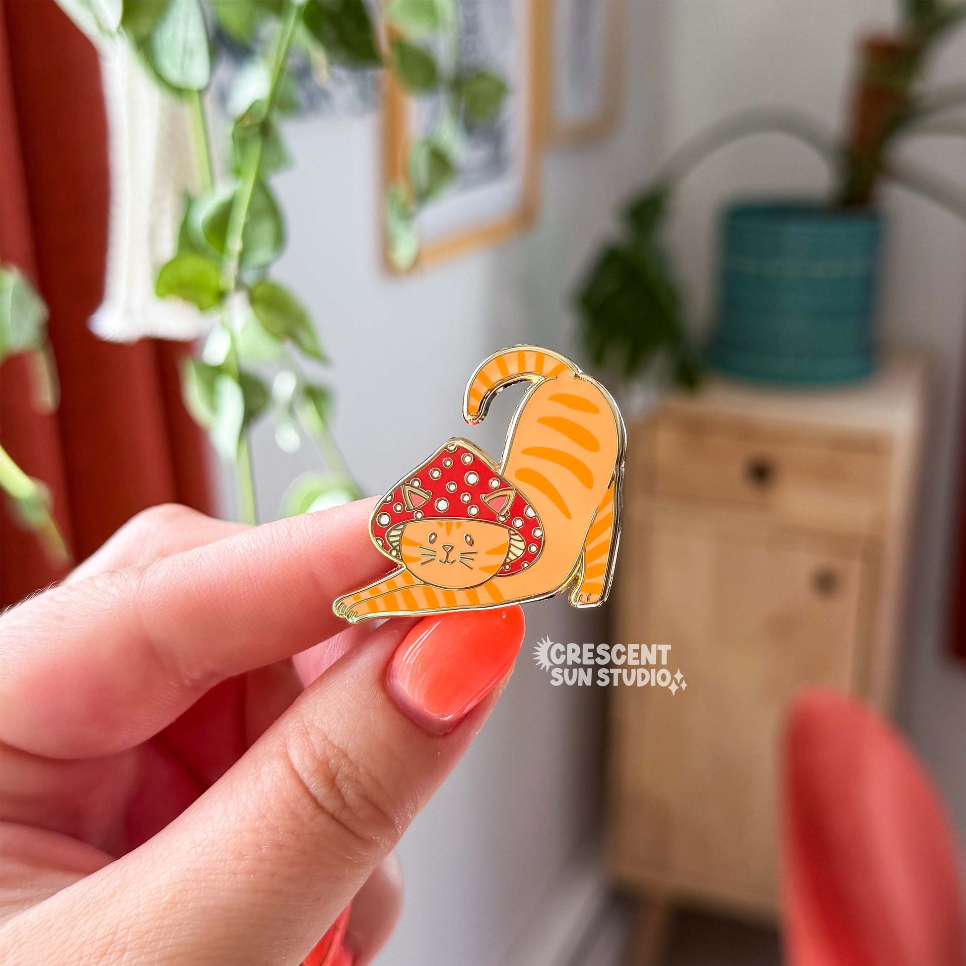 A hand holds an enamel pin in the shape of a cat wearing a mushroom hat is held close to the camera. The cat is a tabby (orange with darker orange stripes) and has a sweet expression on his face. His red mushroom hat with white spots has spots for his ears to poke through. The cat is in a stretching pose with his tail curling up behind him.