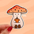 A mushroom shaped sticker of a friendly character wearing an orange mushroom cap and holding a small orange pumpkin