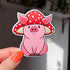 A sticker in the shape of a sitting pig wearing a mushroom hat. This pink shaded pig happily wears a red mushroom with white spots as a hat. His large pig ears stick out of the mushroom.

in this image, a woman's hand holds the sticker close to the camera.