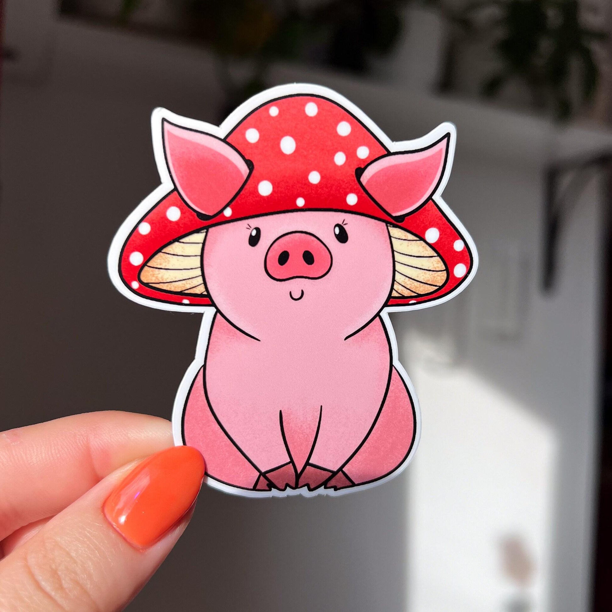 A sticker in the shape of a sitting pig wearing a mushroom hat. This pink shaded pig happily wears a red mushroom with white spots as a hat. His large pig ears stick out of the mushroom.

in this image, a woman's hand holds the sticker close to the camera.