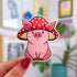 A sticker in the shape of a sitting pig wearing a mushroom hat. This pink shaded pig happily wears a red mushroom with white spots as a hat. His large pig ears stick out of the mushroom.