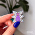 A hand holds a small pin of a purple cat with one large pink eye at the center of its face stoically observes you. The cat has three pairs of light-colored angel wings, each wing covered with watchful pink eyes. 
