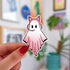 A sticker in the shape of a ghost wearing a pink cat ear headseat. Her body is in gradient pink and you can tell she is involved in an intense conversation