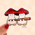 Die cut sticker of two plump beige mushroom characters with red and white spotted caps, sit side by side sharing a lit blunt.