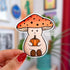 A mushroom shaped sticker of a friendly character wearing an orange mushroom cap and holding a small orange pumpkin