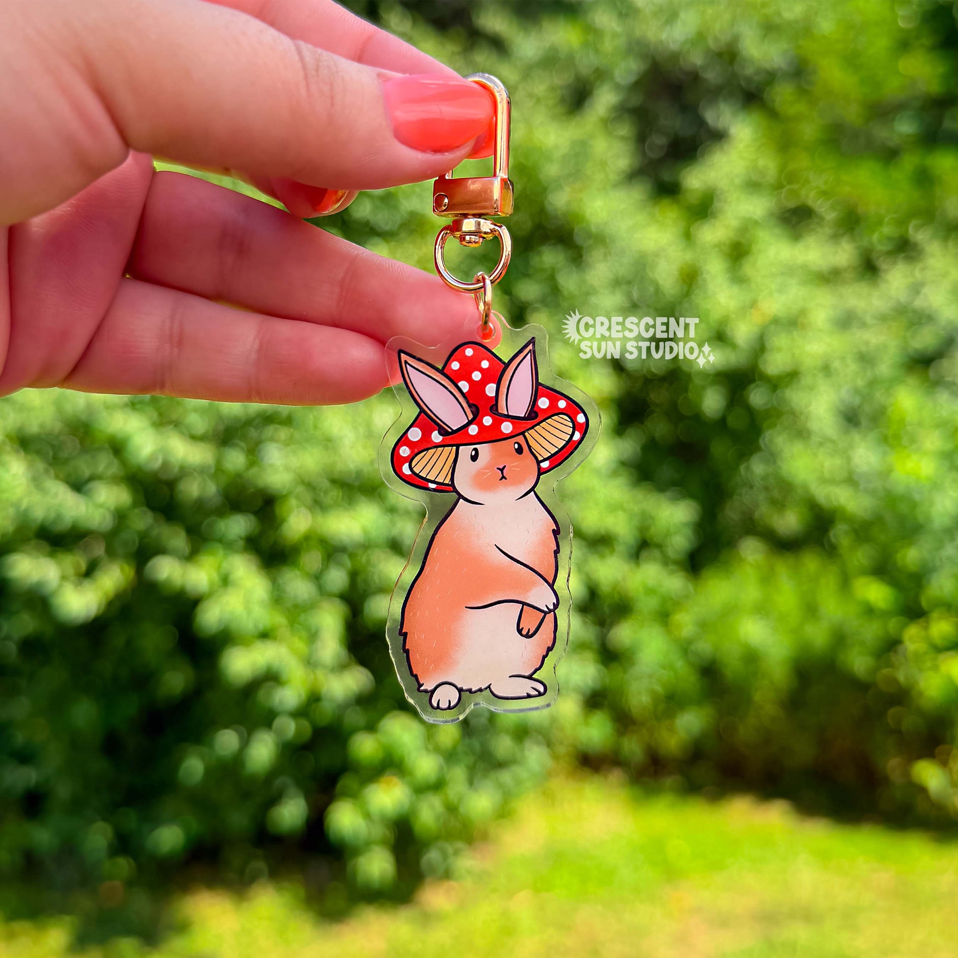 Acrylic die cut of a fluffy brown and white rabbit wearing a red spotted mushroom cap, standing upright with a composed expression. Keychain has a gold u-shaped clasp.
