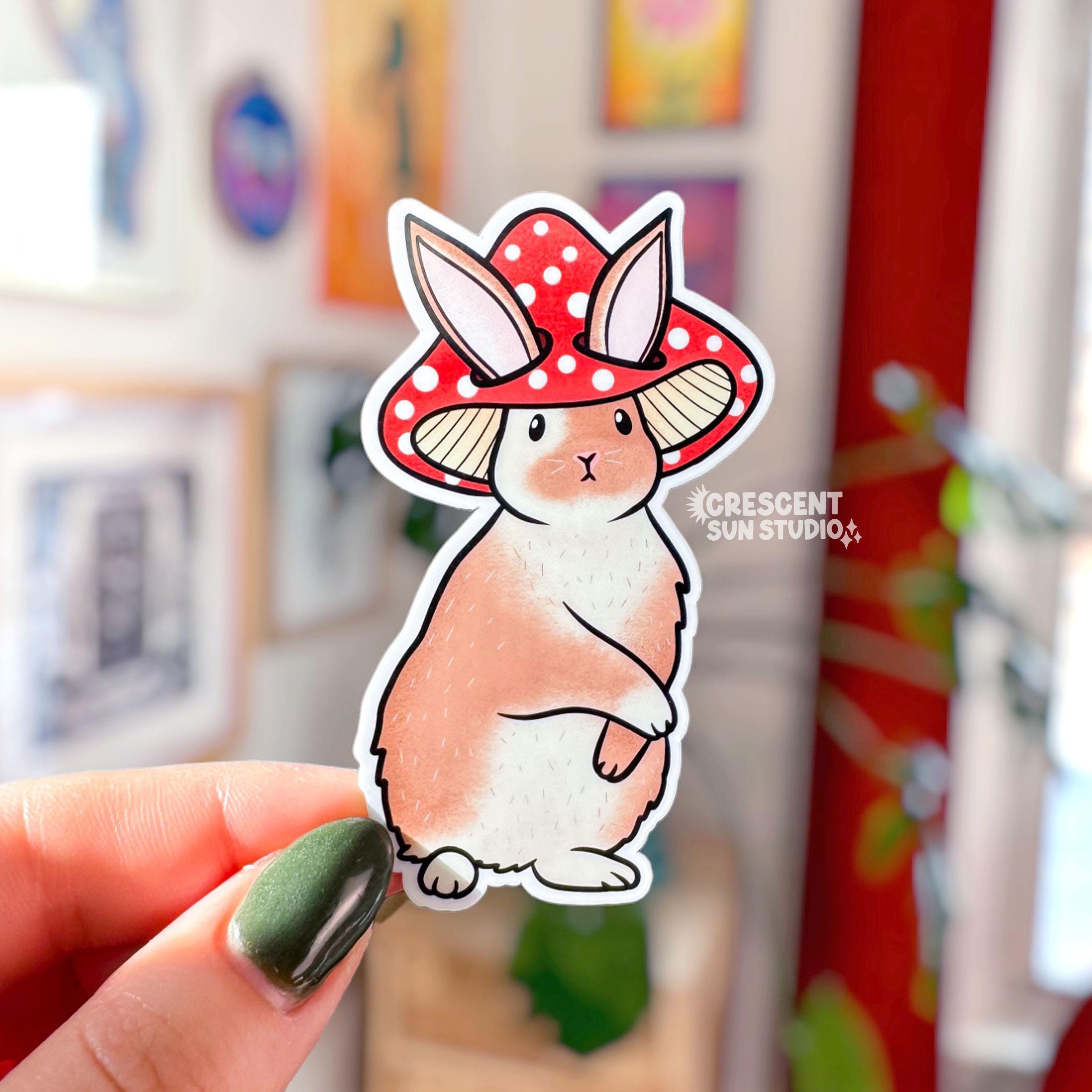 Die-cut sticker of a fluffy brown and white rabbit wearing a red spotted mushroom cap, standing upright with a composed expression.