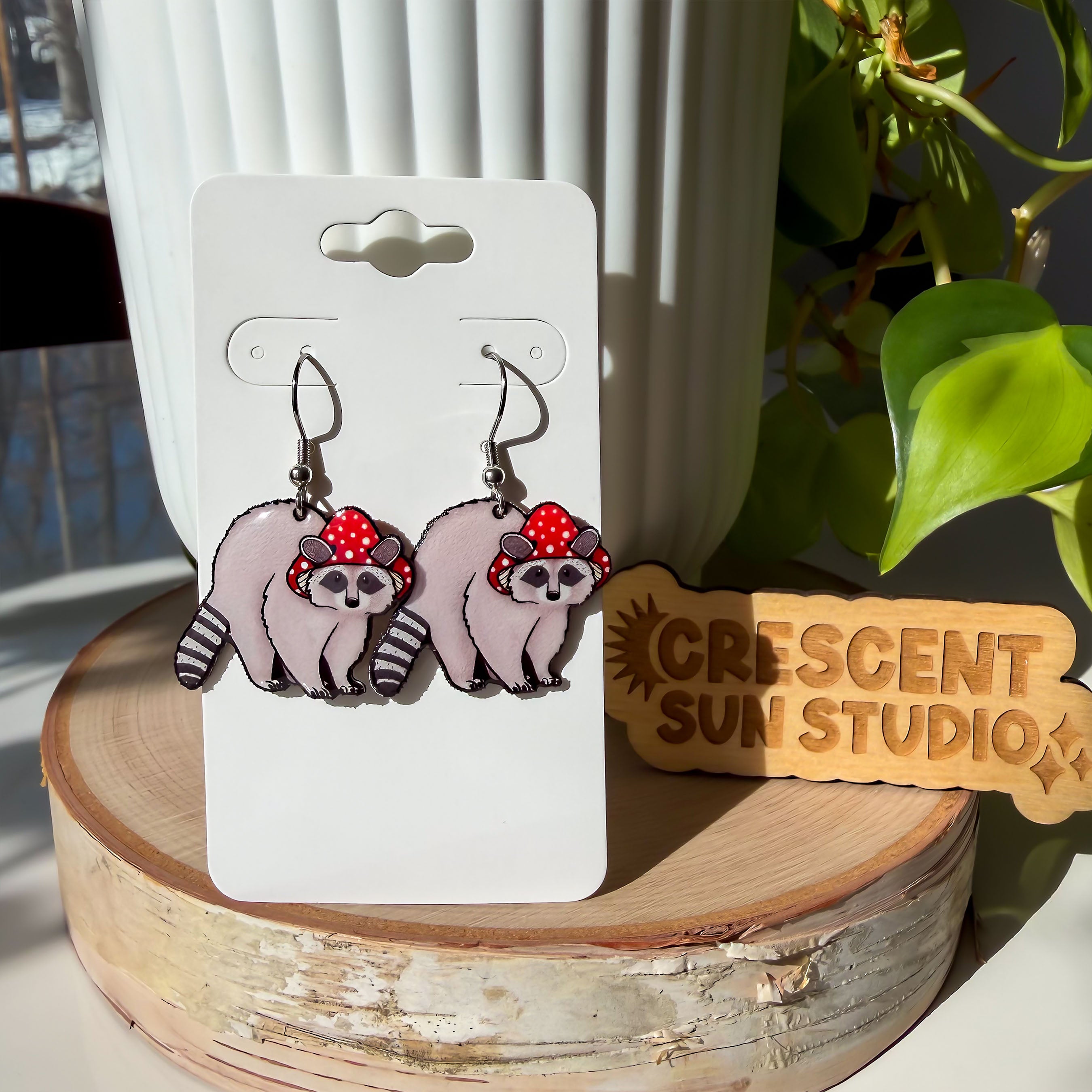 Dangle earrings featuring a gray raccoon with a striped tail. The raccoon is wearing a red mushroom cap with white spots. The cap has little holes for the raccoon's ears to poke through.

The earrings are displayed on a white card that is propped up against a white plant pot.