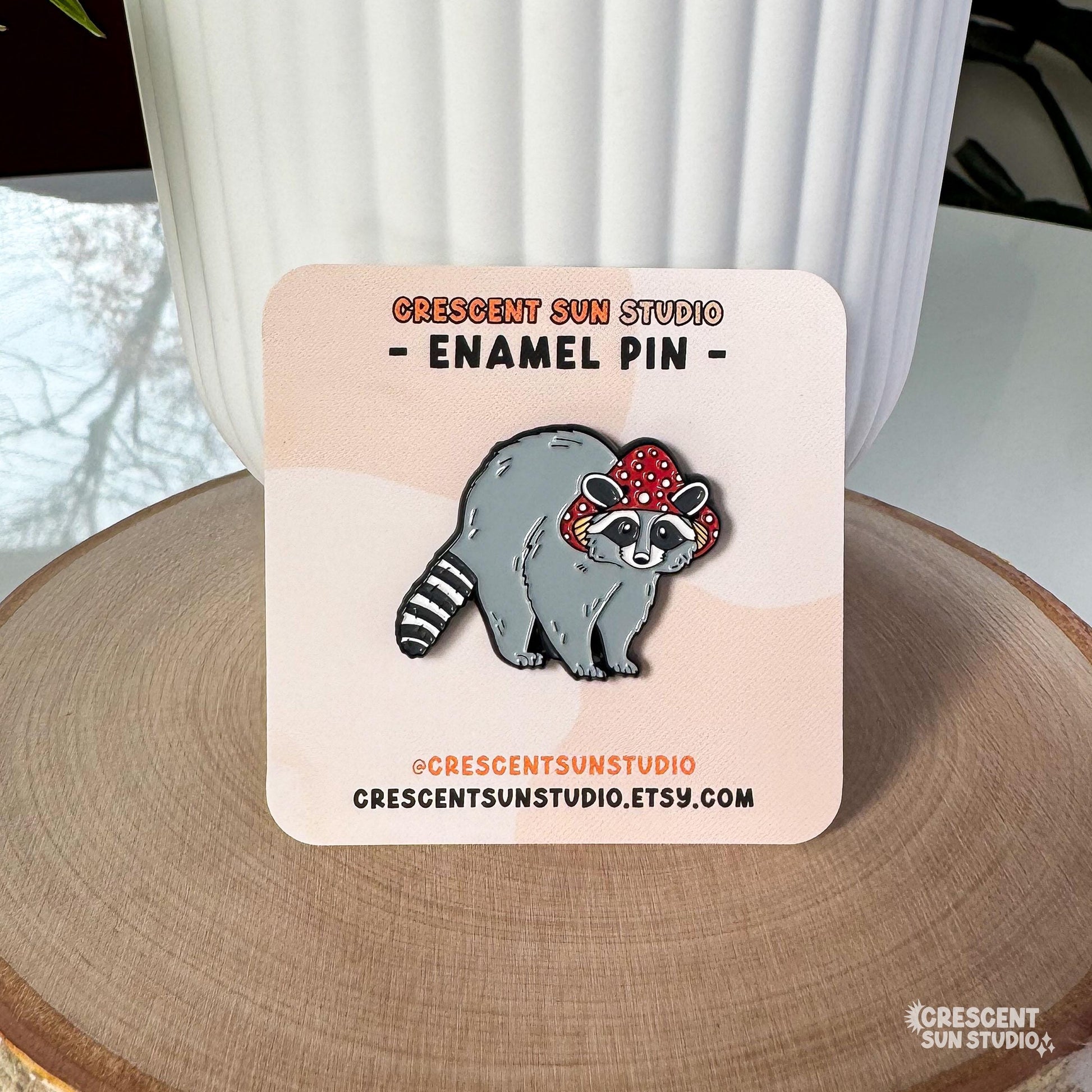 Displayed on a light colored card, this enamel pin features a gray raccoon with a striped tail. The raccoon is wearing a red mushroom cap with white spots. The cap has little holes for the raccoon's ears to poke through.
