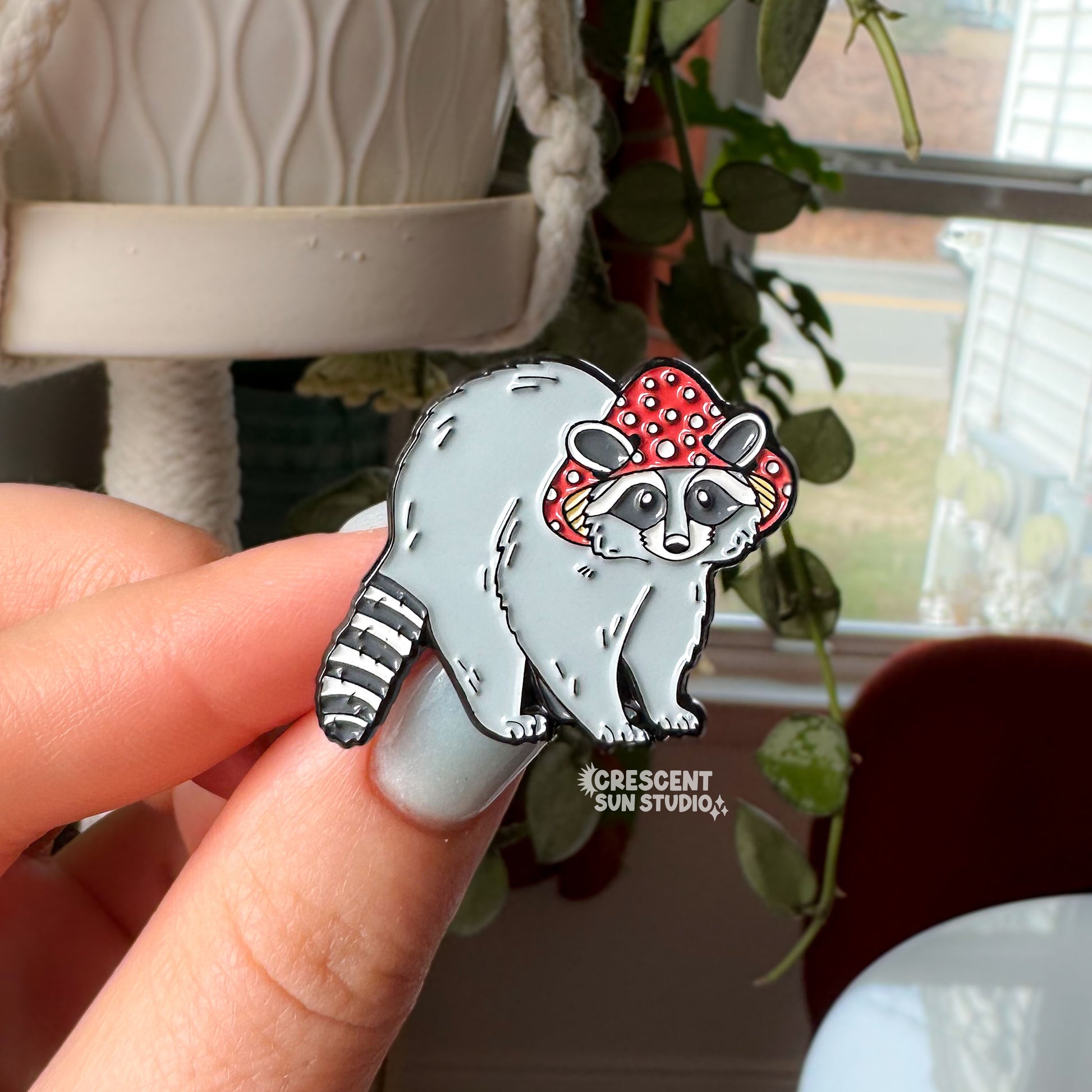 This enamel pin features a gray raccoon with a striped tail. The raccoon is wearing a red mushroom cap with white spots. The cap has little holes for the raccoon's ears to poke through.

In this image, the pin is being held close to the camera by a person's hand.