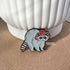 Shown out of the packaging and propped up against a white plant pot, this enamel pin features a gray raccoon with a striped tail. The raccoon is wearing a red mushroom cap with white spots. The cap has little holes for the raccoon's ears to poke through.
