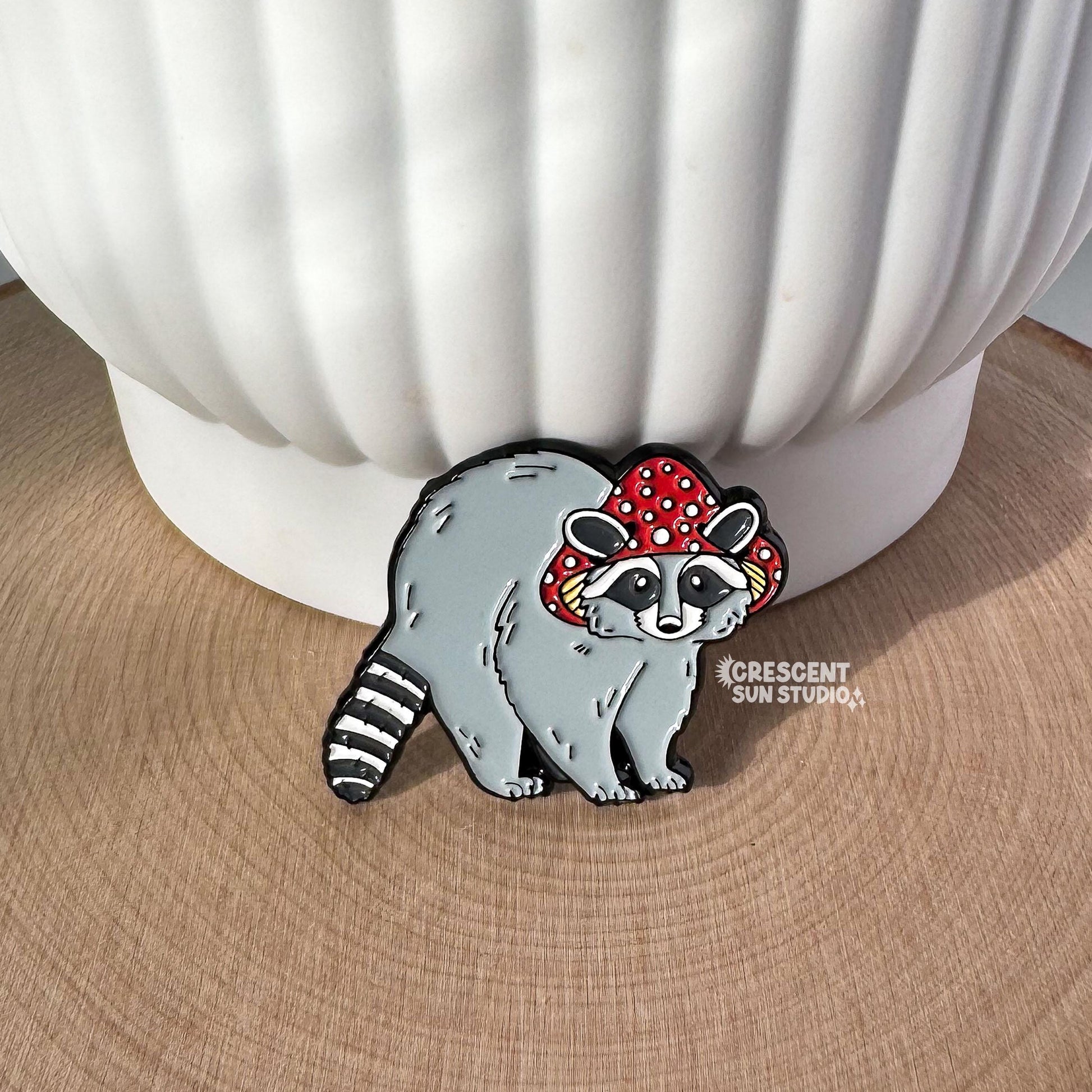 Shown out of the packaging and propped up against a white plant pot, this enamel pin features a gray raccoon with a striped tail. The raccoon is wearing a red mushroom cap with white spots. The cap has little holes for the raccoon's ears to poke through.
