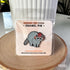 Displayed on a light colored card and enclosed in clear plastic packaging, this enamel pin features a gray raccoon with a striped tail. The raccoon is wearing a red mushroom cap with white spots. The cap has little holes for the raccoon's ears to poke through.

The pin and its packaging are propped up on a wood stand and lean against a white pot.
