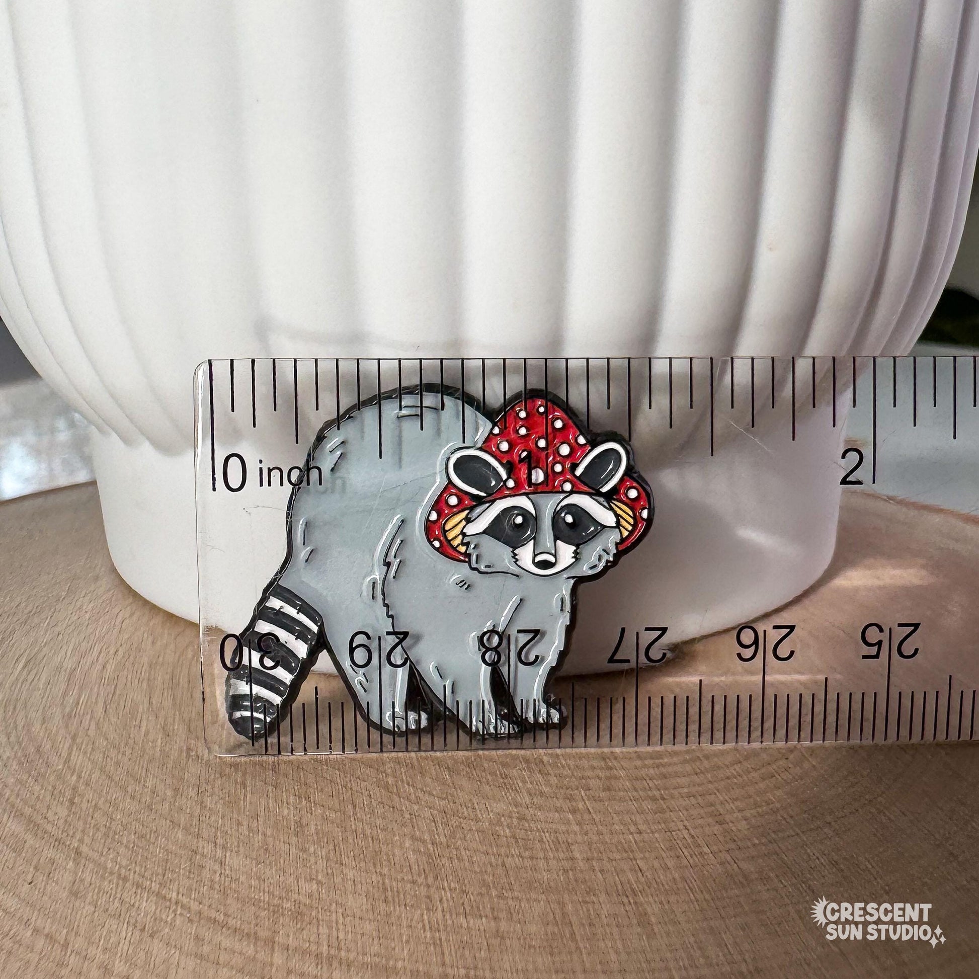 This enamel pin features a gray raccoon with a striped tail. The raccoon is wearing a red mushroom cap with white spots. The cap has little holes for the raccoon's ears to poke through.

A clear plastic ruler sits in front of the pin, showing its size of 1.35 inches.
