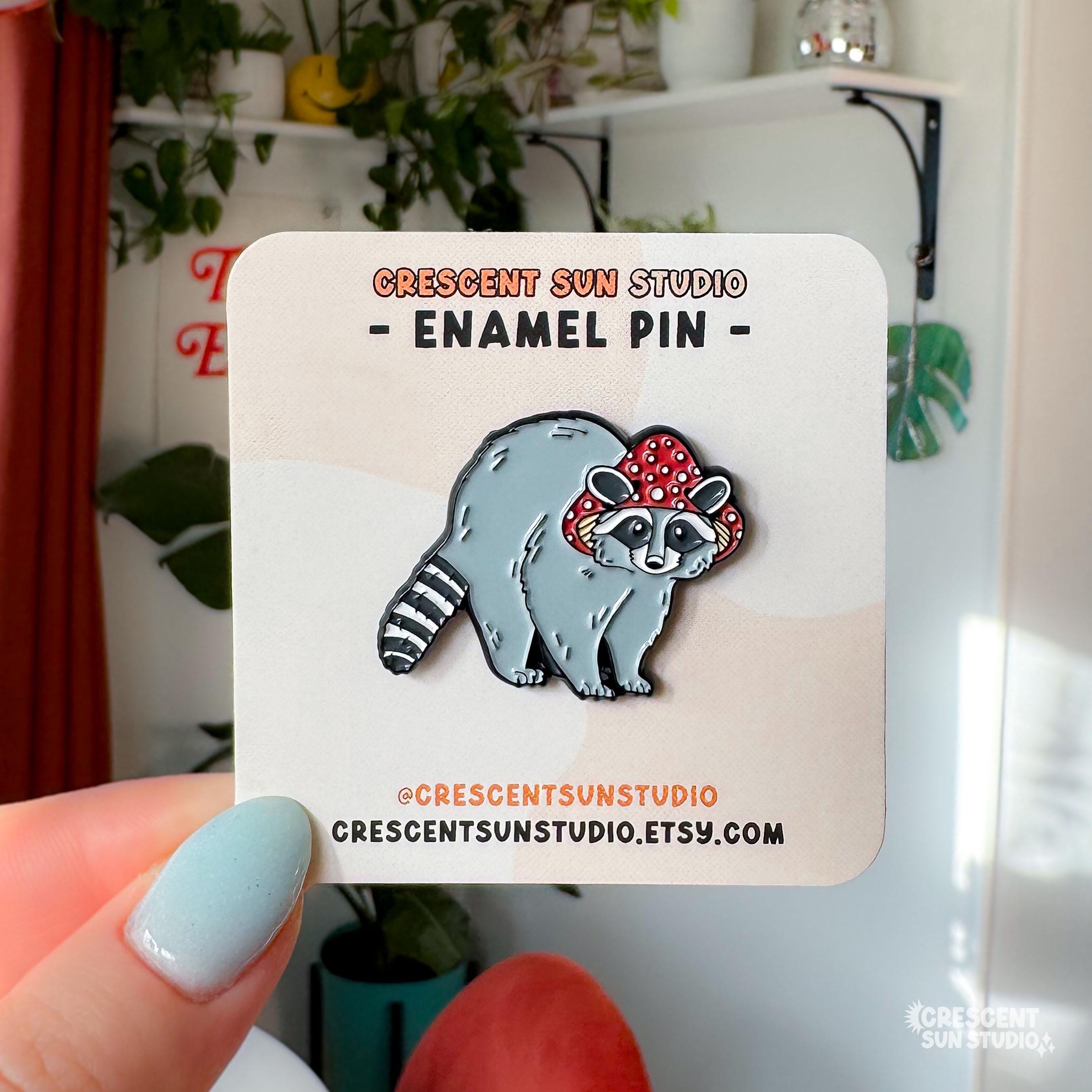 Displayed on a light colored card, this enamel pin features a gray raccoon with a striped tail. The raccoon is wearing a red mushroom cap with white spots. The cap has little holes for the raccoon's ears to poke through.

The card is held by a person's hand and her pastel blue colored thumb is visible in the photo.