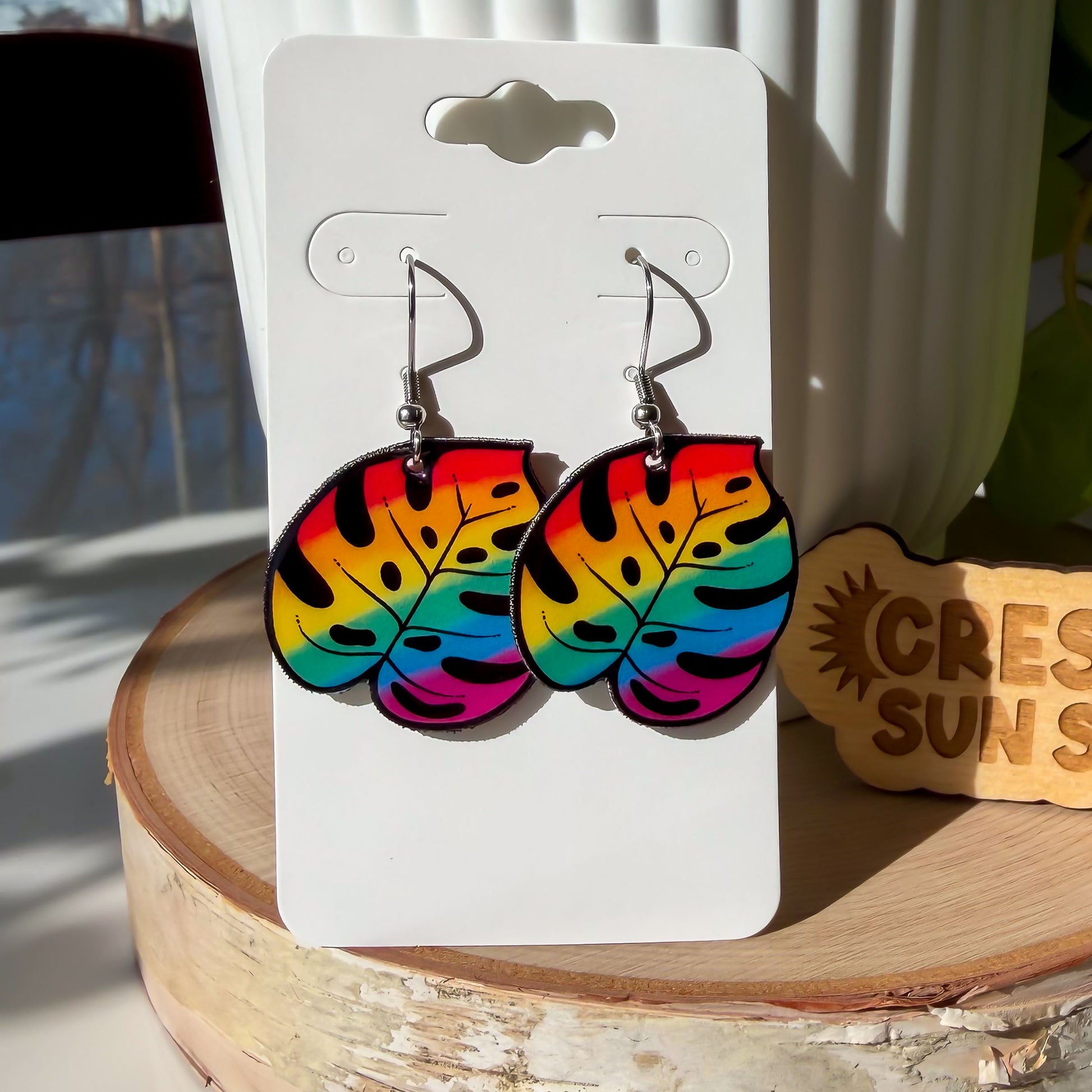 A close up of our shrink plastic dangle earrings shaped like monstera leaves, printed in a bold rainbow gradient from red and orange at the top to teal and magenta at the bottom, with black cutout details.

These earrings are displayed on a white card, which is propped up against a white plant pot.