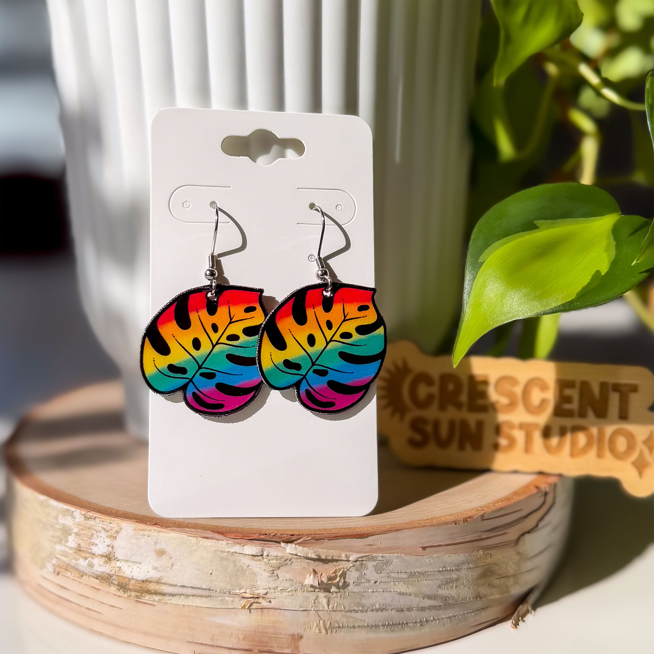 Shrink plastic dangle earrings shaped like monstera leaves, printed in a bold rainbow gradient from red and orange at the top to teal and magenta at the bottom, with black cutout details.

These earrings are displayed on a white card, which is propped up on a white plant pot. Everything sits on a wooden stand