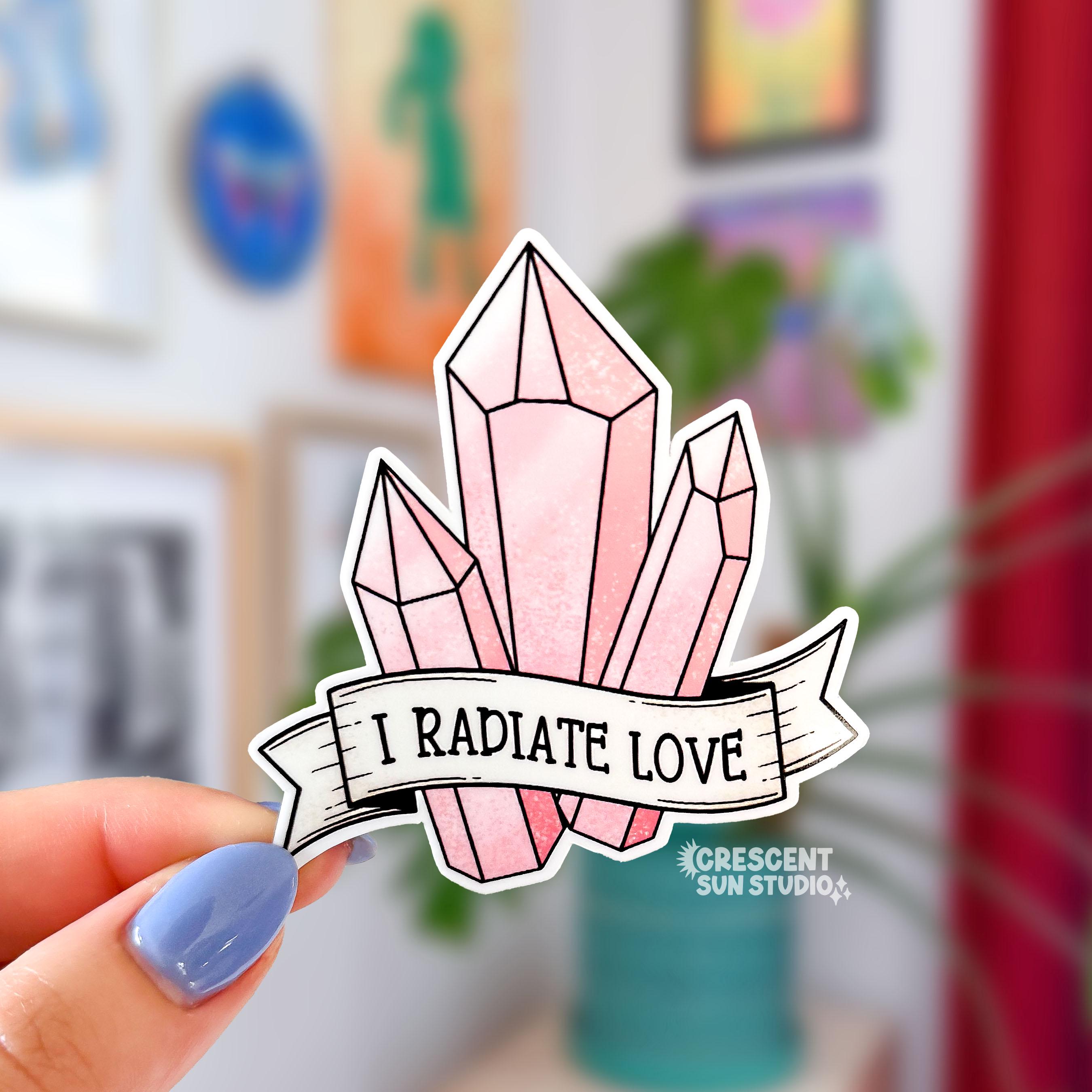 A sticker in the shape of three pink shaded crystals. At the bottom is a white banner with black text that reads "I radiate love"