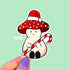 A die-cut sticker of a mushroom wearing a santa hat on top of his red and white spotted cap. The mushroom is a pinky-beige color, has a smile on his face, and his holding a giant candy cane.