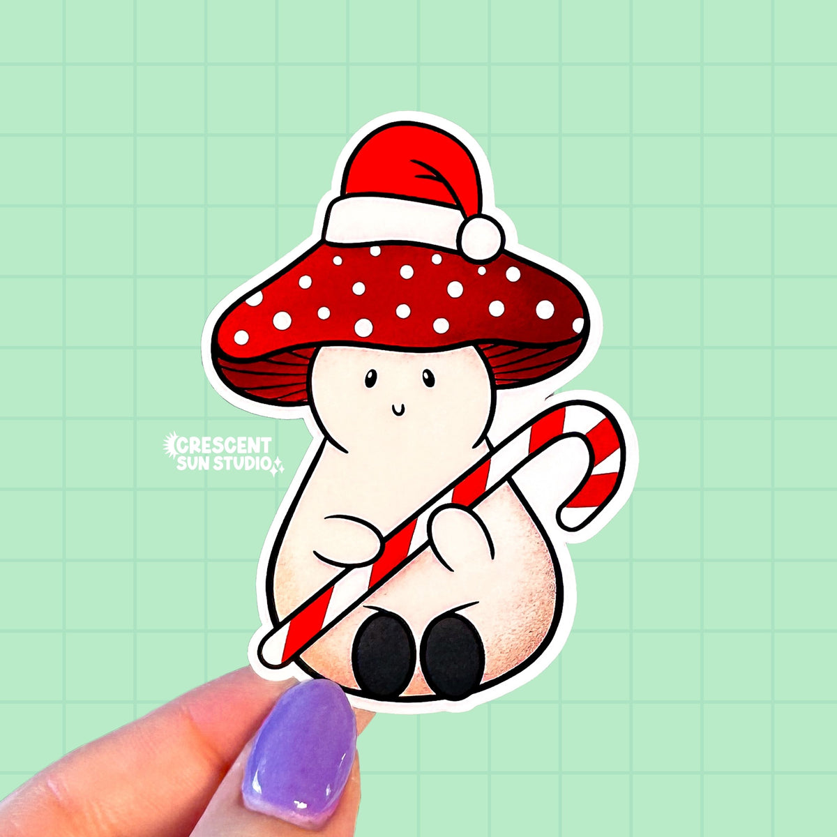A die-cut sticker of a mushroom wearing a santa hat on top of his red and white spotted cap. The mushroom is a pinky-beige color, has a smile on his face, and his holding a giant candy cane.