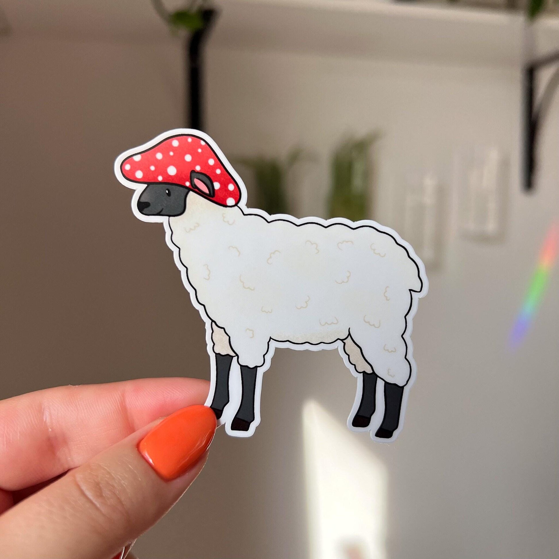 A die-cut sticker of a white wooly sheep with a black muzzle and legs. The sheep wears a red mushroom with white spots for a hat. The sheep's ears poke out through the hat.

In this image, the sticker is held close to the camera by a woman's hand.