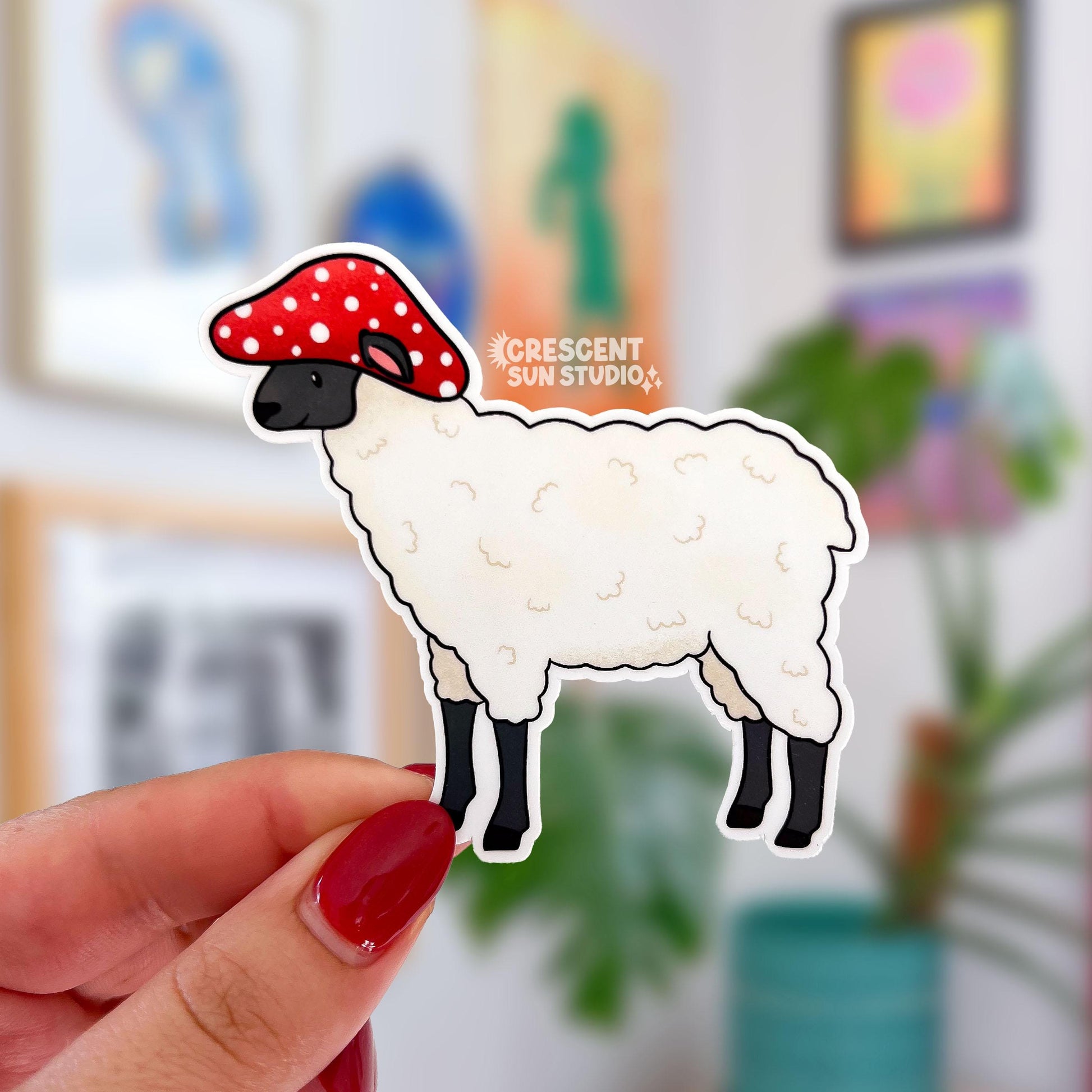 A die-cut sticker of a white wooly sheep with a black muzzle and legs. The sheep wears a red mushroom with white spots for a hat. The sheep's ears poke out through the hat.