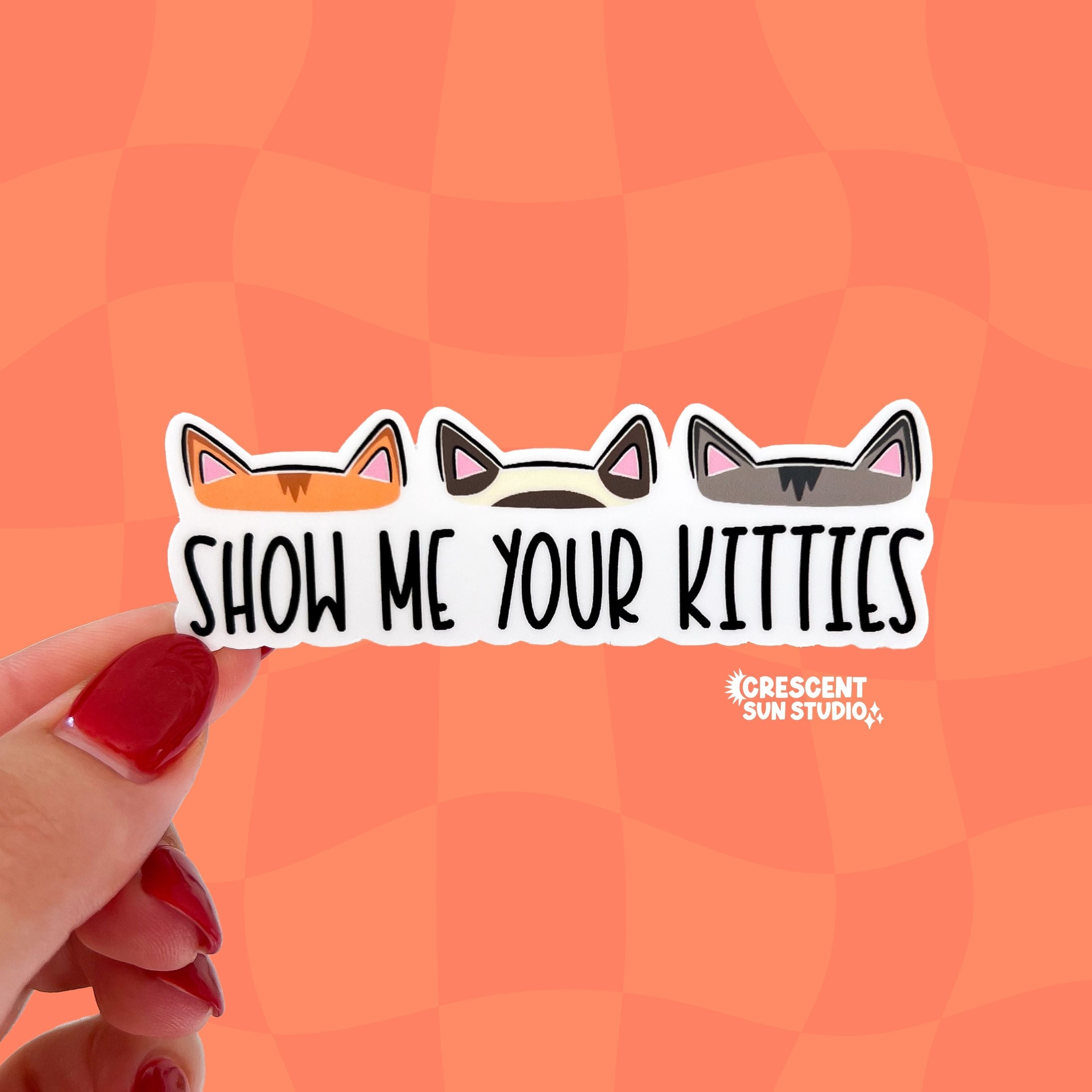This rectangle-shaped sticker features the text "show me your kitties." Above the text are three different pairs of cat ears - one set is orange, the other is cream and brown, and the last set of kitty ears is a medium grey. The sticker is cut to reflect the shape of each individual pair of cat ears.