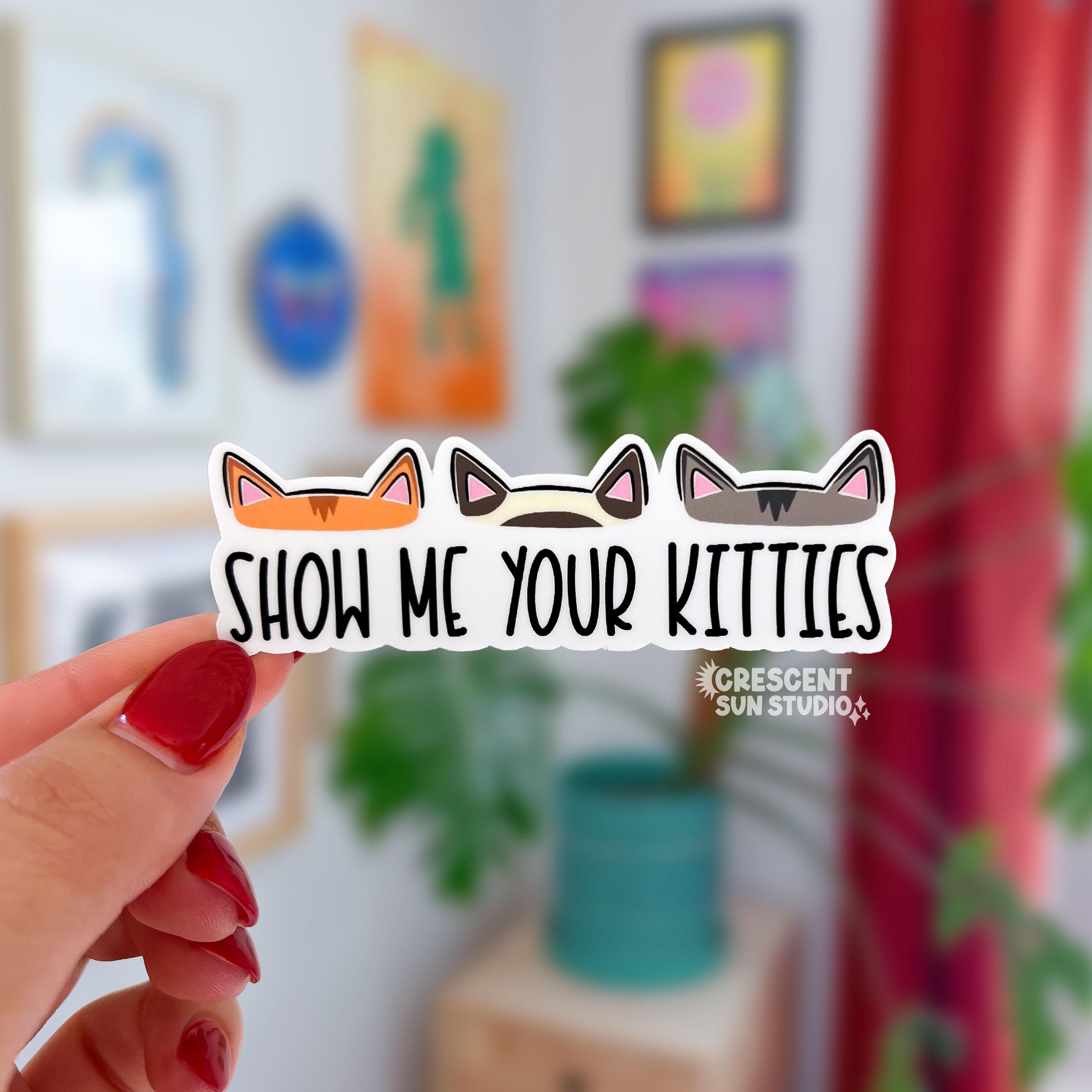 This rectangle-shaped sticker features the text "show me your kitties." Above the text are three different pairs of cat ears - one set is orange, the other is cream and brown, and the last set of kitty ears is a medium grey. The sticker is cut to reflect the shape of each individual pair of cat ears.