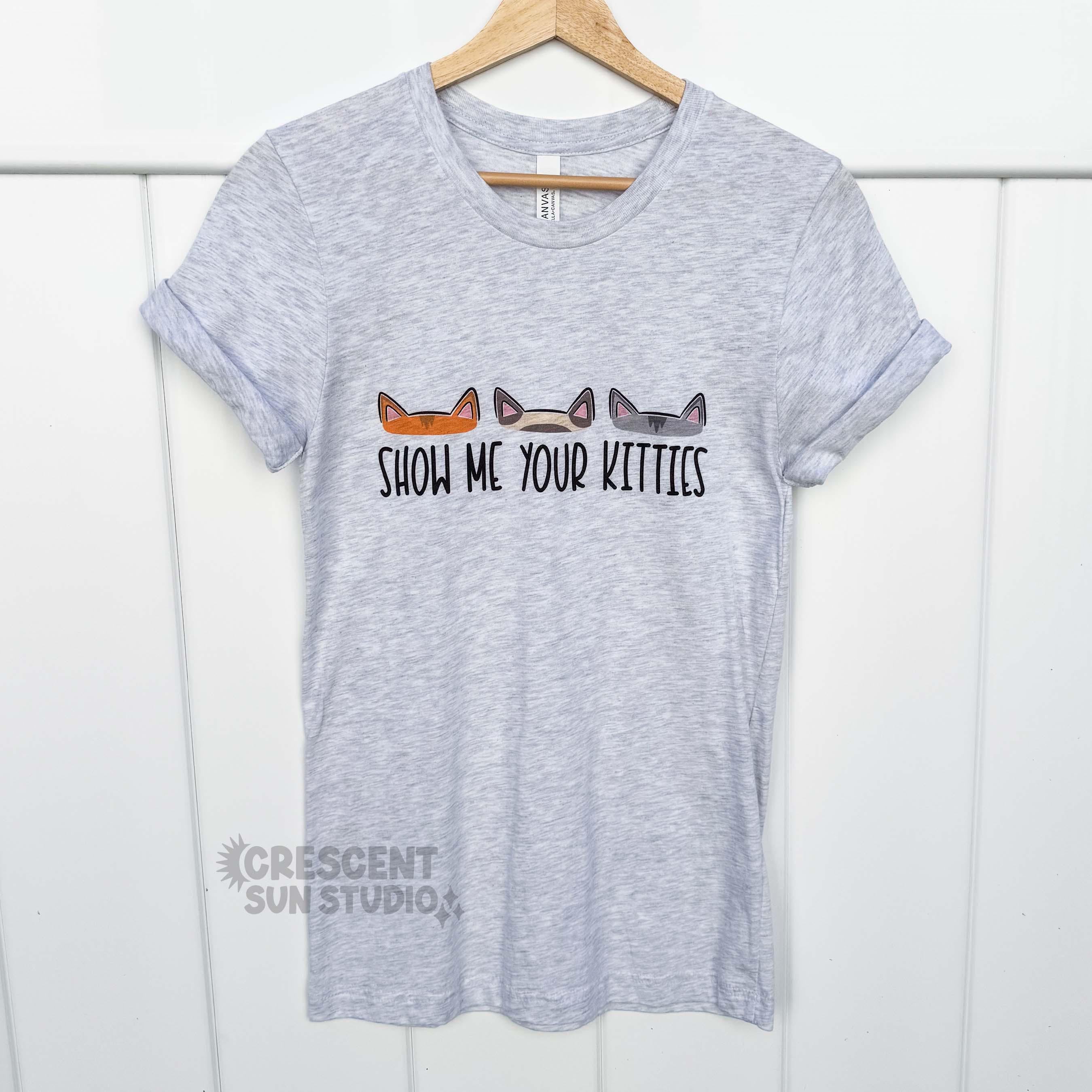 A light heather grey t-shirt hangs on a hanger. On the shirt is the text "show me your kitties." Above the text are three different pairs of cat ears - one set is orange, the other is cream and brown, and the last set of kitty ears is a medium grey.