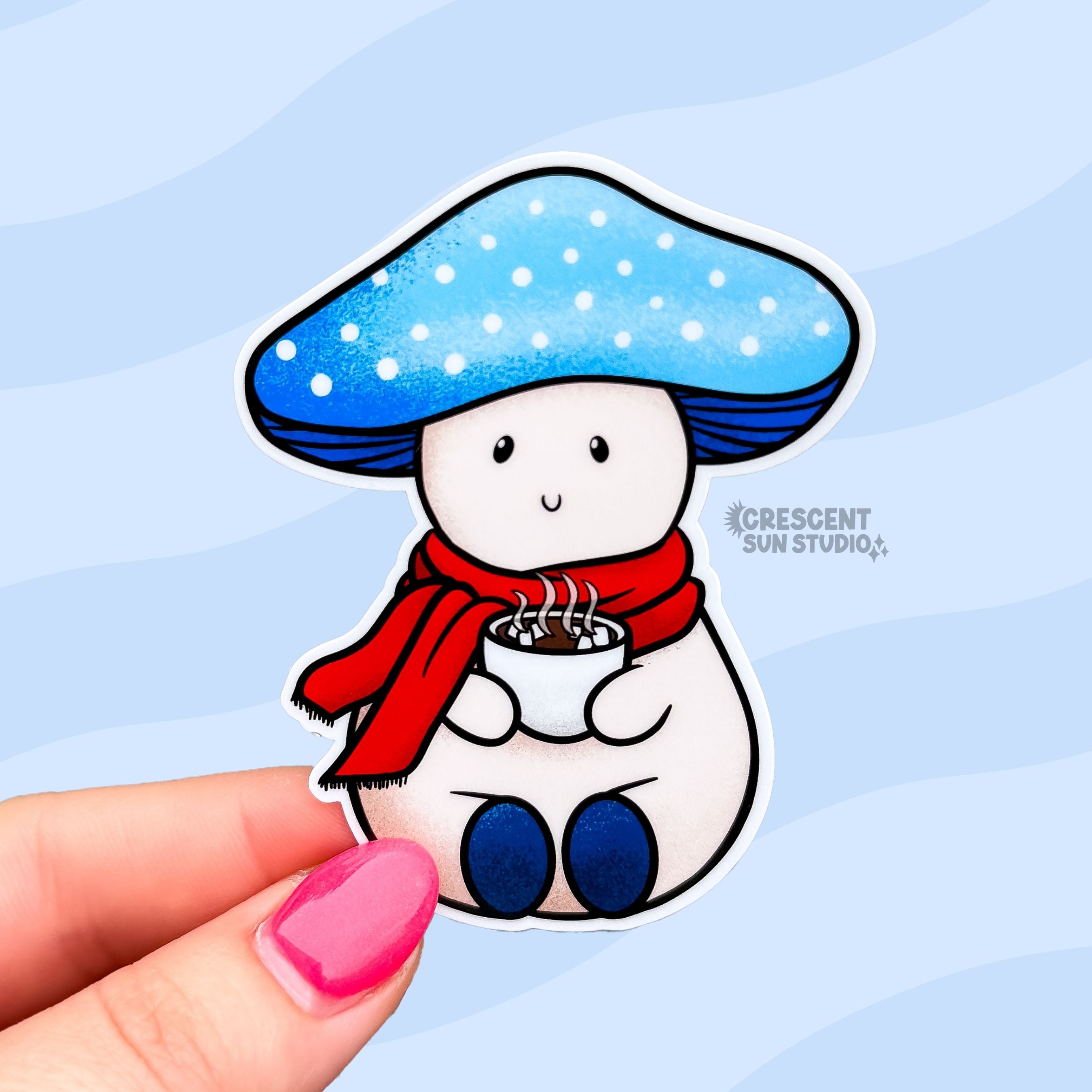 A die-cut sticker of a light colored mushroom with a light blue mushroom cap. The cap has white dots on it and Snowy has a content expression. He is wearing a red scarf and holding a steaming cup of hot cocoa.