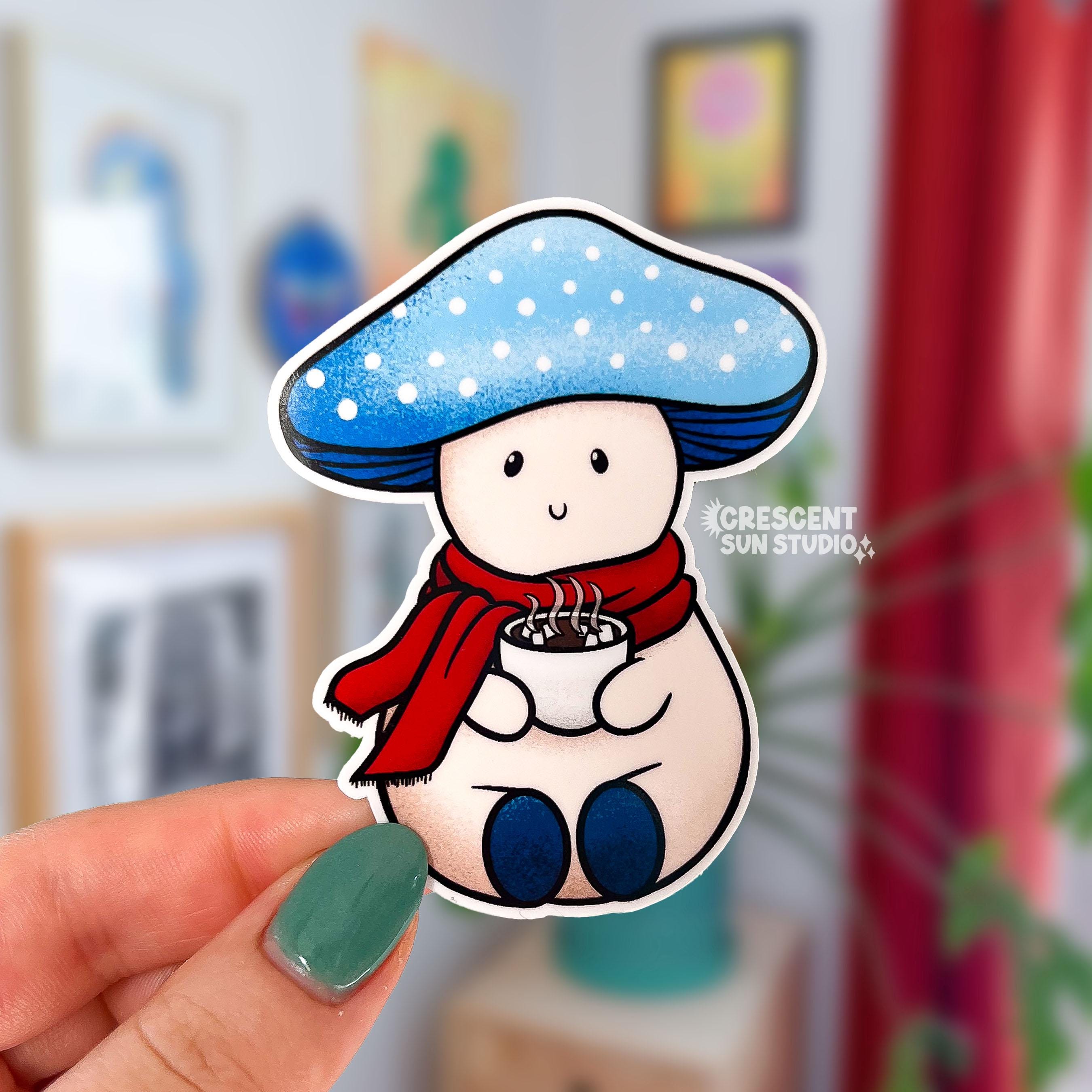 A die-cut sticker of a light colored mushroom with a light blue mushroom cap. The cap has white dots on it and Snowy has a content expression. He is wearing a red scarf and holding a steaming cup of hot cocoa.