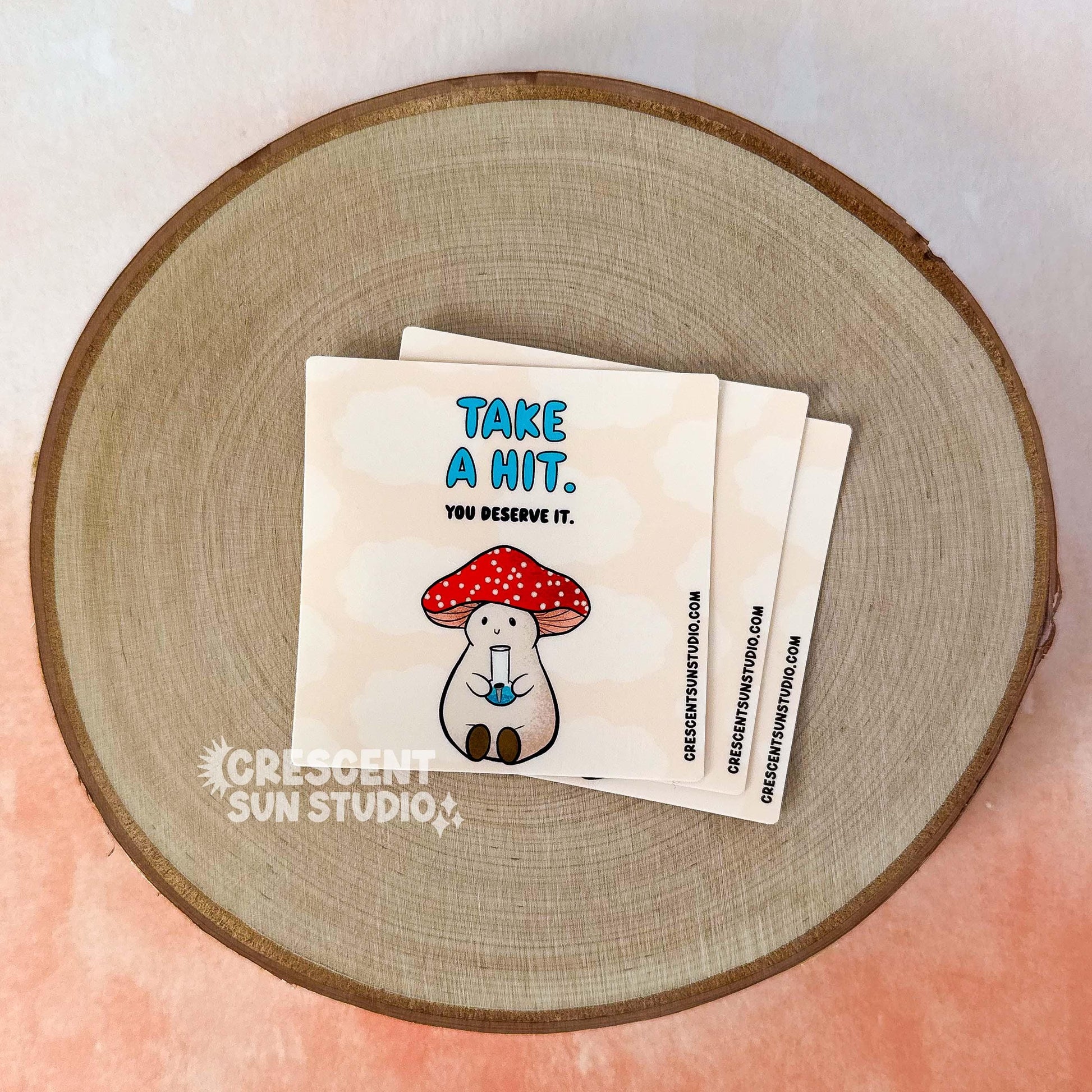 A stack of lighter wraps: each with a peachy-beige background.  On the top of the wrap in blue letters it reads, "take a hit" and below that it says "you deserve it." At the bottom of the wrap sits Stoney the Mushroom, our pinky-beige plump mushroom character with a red and white spotted cap is joyfully holding a bong in his little mushroom hands.