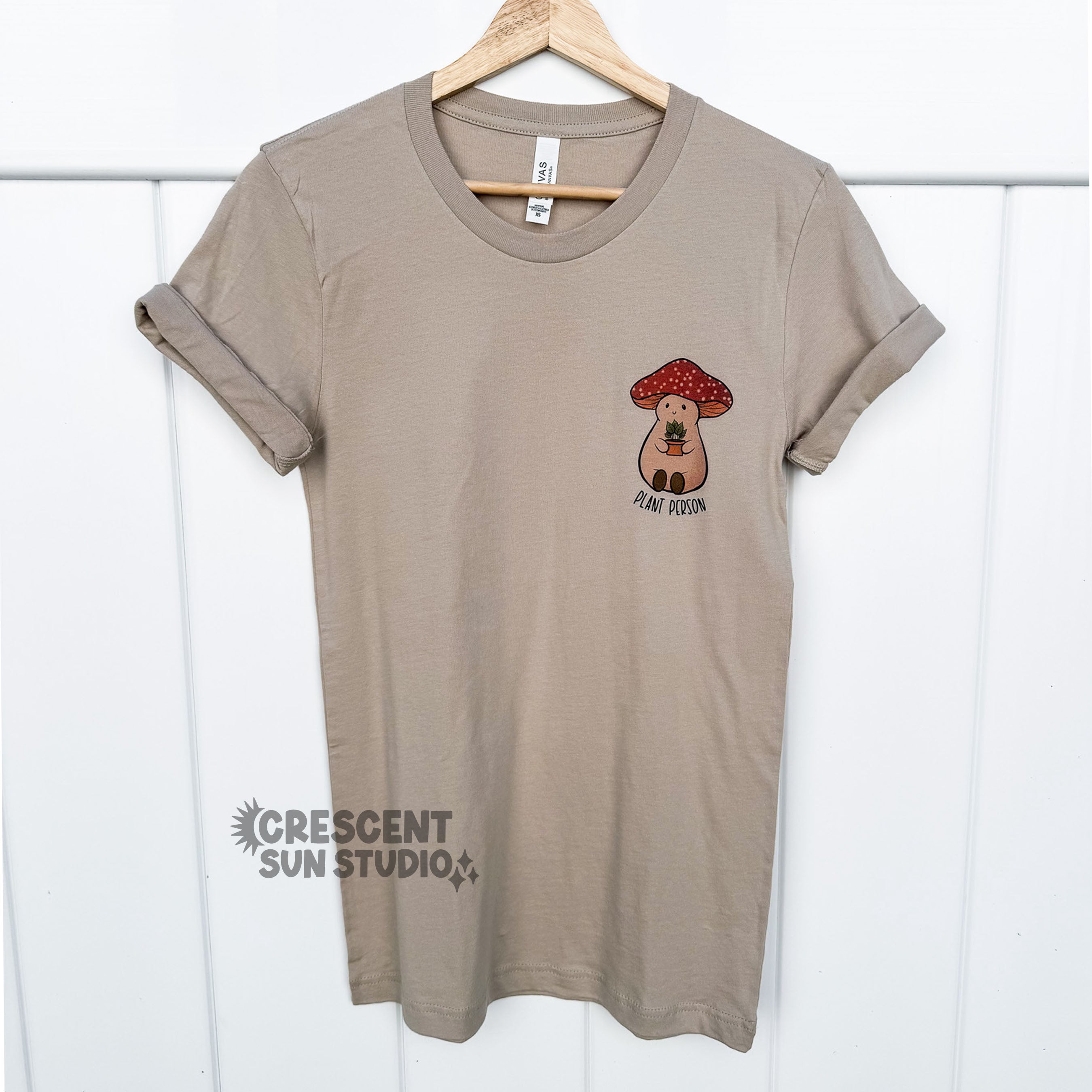 A tan colored shirt hangs on a wooden hanger against a white wall. On the left chest area of the shirt (right in the photograph) is our Stoney the Mushroom design. He is a plump, smiling pinky-beige mushroom character with a red mushroom cap with white spots. Stoney is holding a tiny potted plant in his little mushroom arms. 

Underneath Stoney is the text "plant person" written in black.