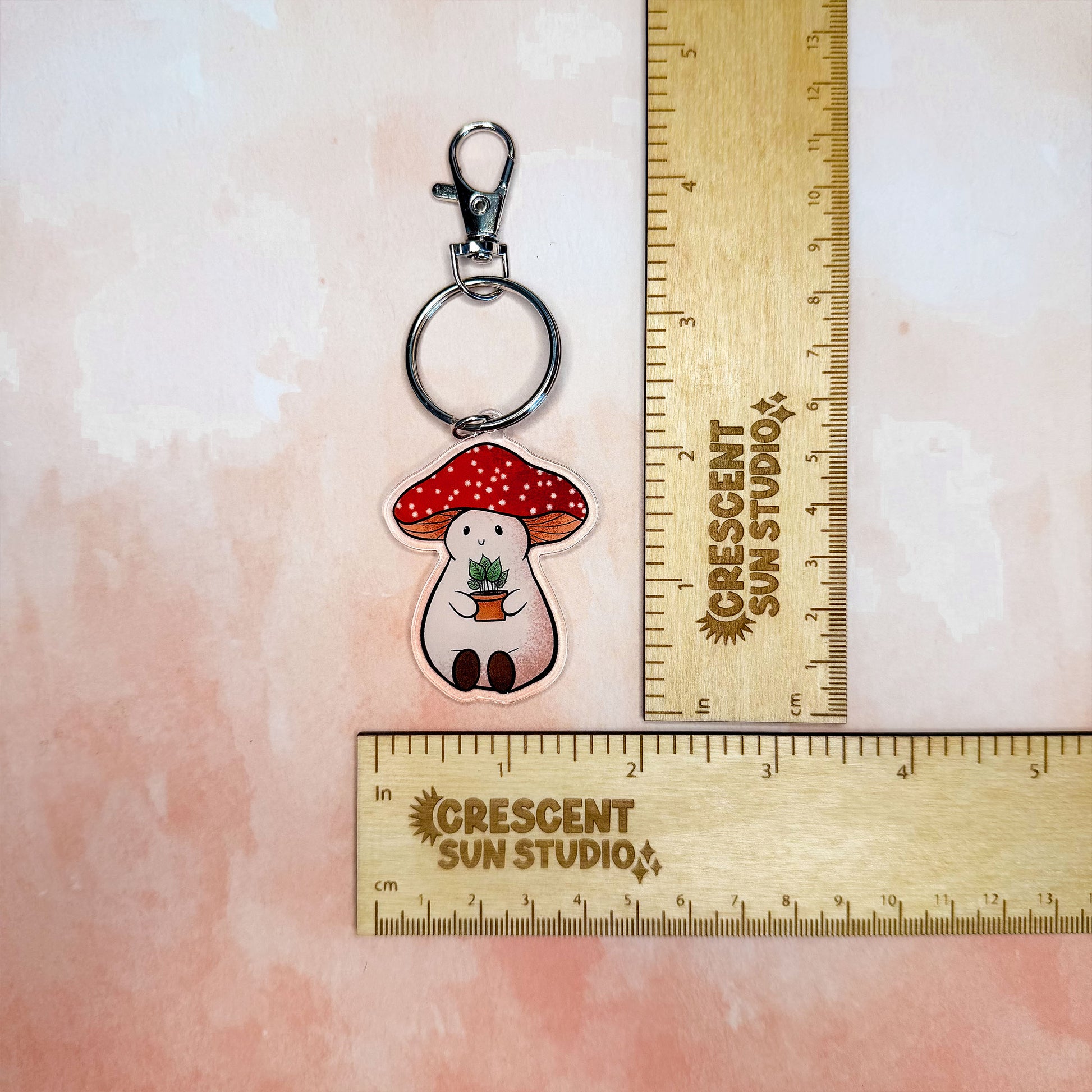 In this image, the Stoney keychain sits next to to rulers, one placed vertically and one placed horizontally, showing Stoney's full size, including the keychain ring and clasp, which is roughly 4.5 x 1.6 inches.

Stoney the Mushroom is a plump, smiling pinky-beige mushroom character with a red mushroom cap with white spots. Stoney is holding a tiny potted plant in his little mushroom arms. 